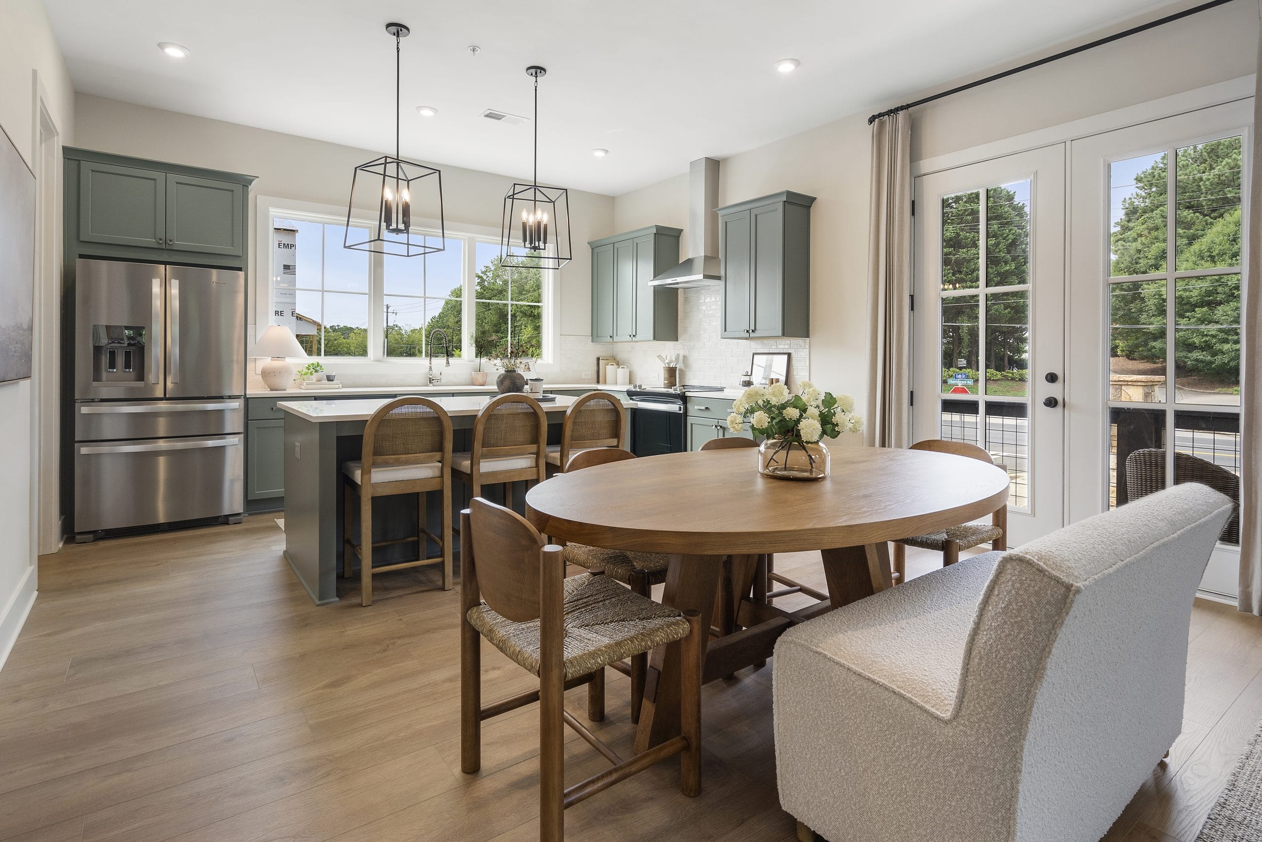 Kitchen and dining area with green cabinetry, stainless steel appliances, central island with wooden bar stools, round wooden dining table, large windows, and glass doors.