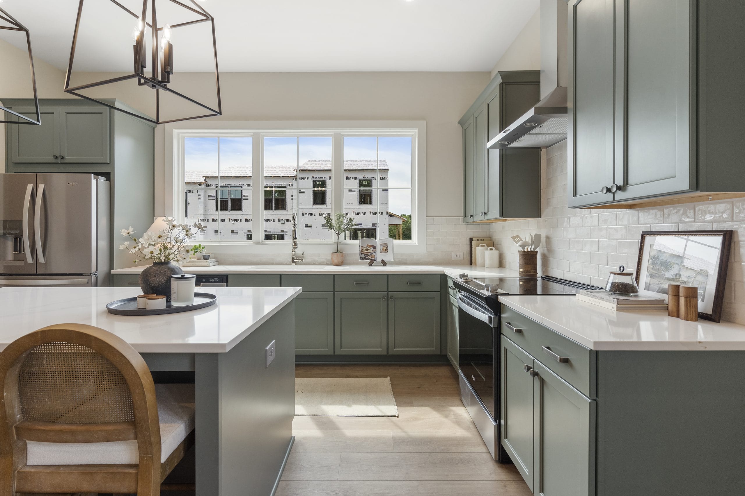 Kitchen with green cabinetry, white countertops, stainless steel appliances, large window, kitchen island with wooden chair, white subway tile backsplash, and pendant lights.