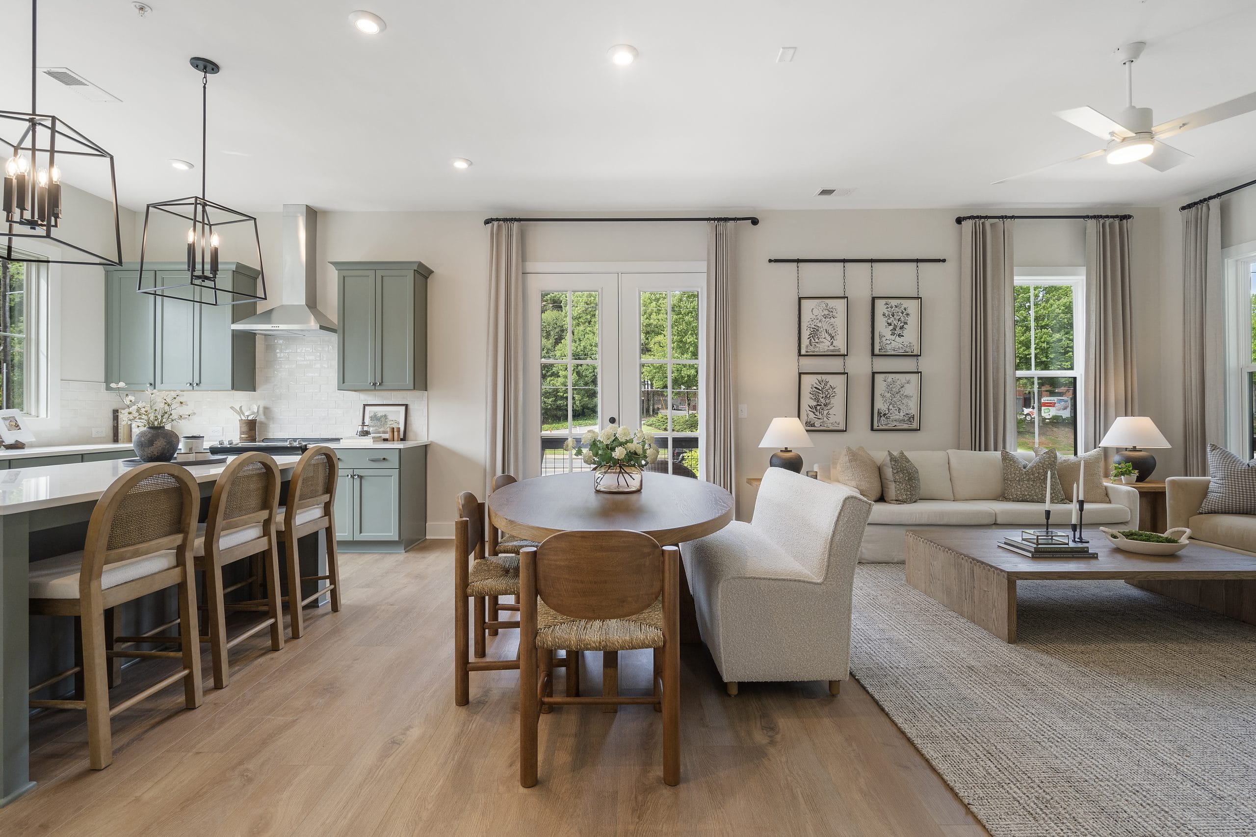 Living and dining area with light green kitchen cabinets, stainless steel range hood, beige sectional sofa, large windows, and botanical prints