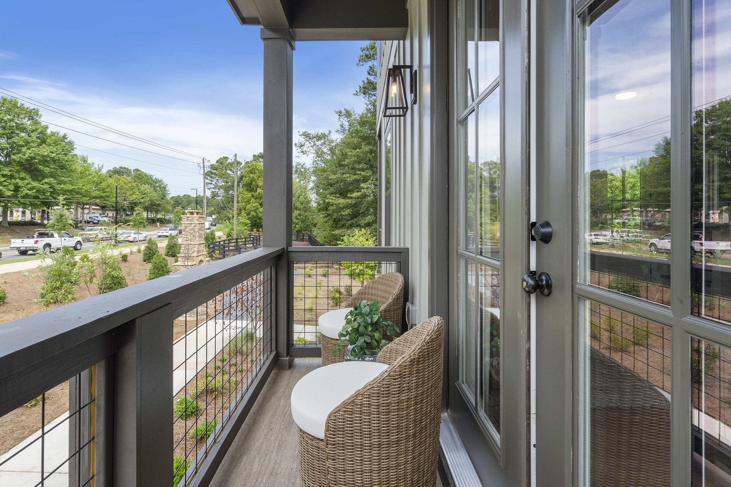 Small balcony with wicker chairs, round table with potted plant, wire mesh railing, view of trees, road, and buildings.