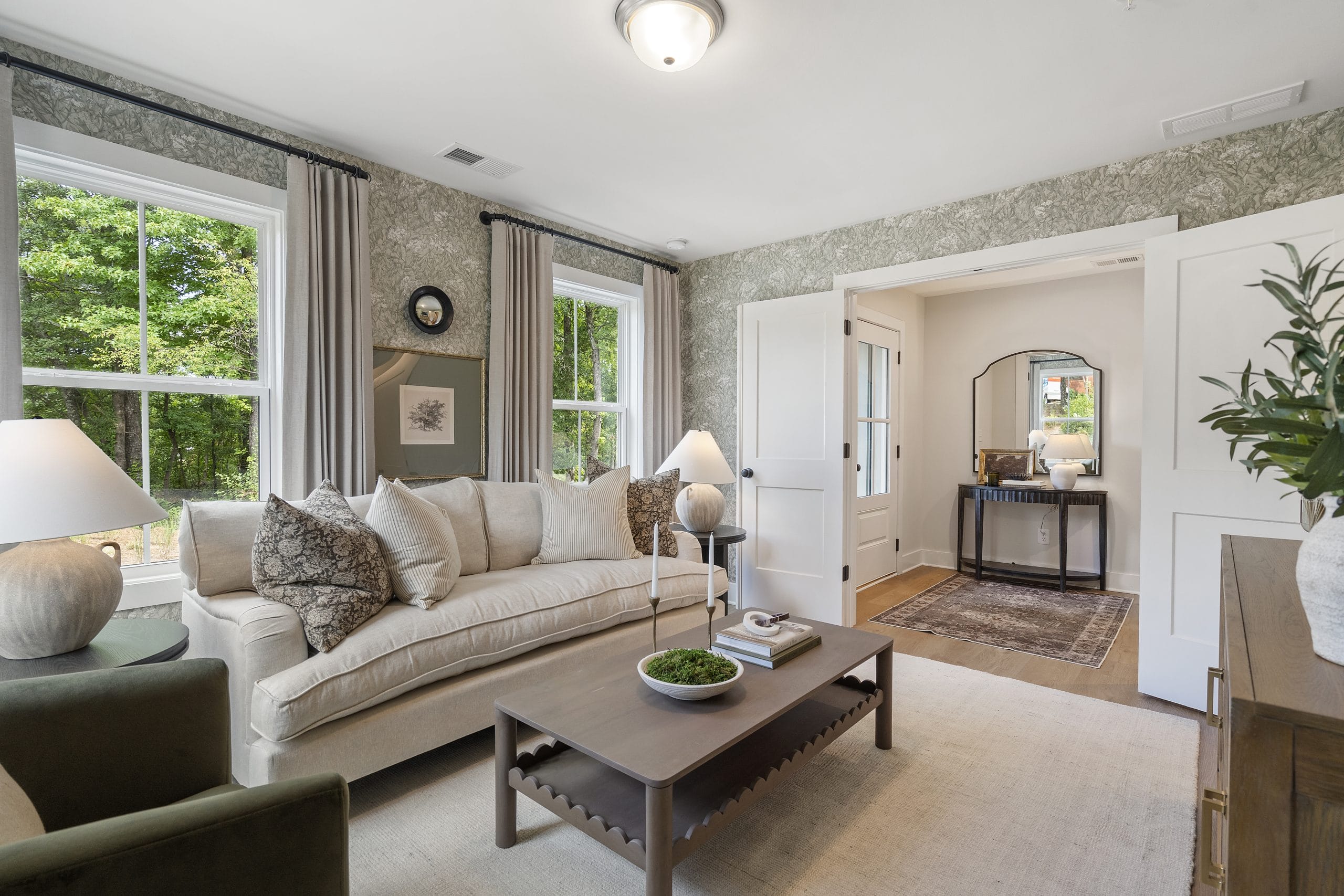 Living room with beige sofa, wooden coffee table with decorative items, armchair, large windows with curtains, framed pictures, patterned wallpaper, and overhead light fixture.