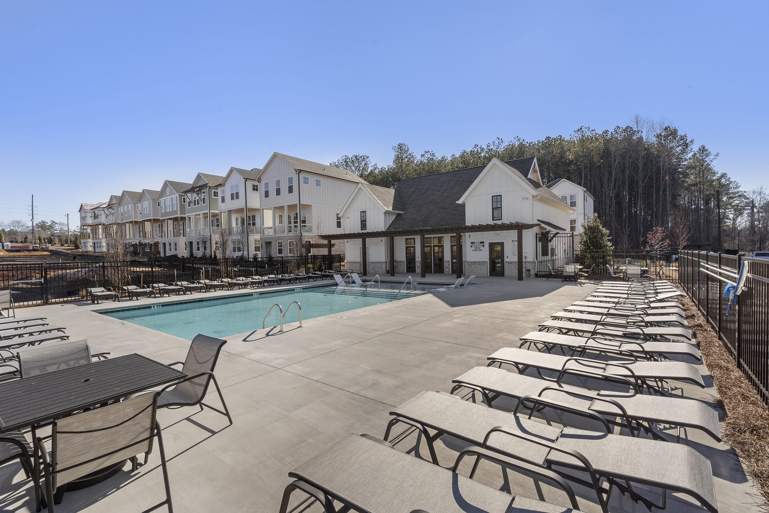 Outdoor swimming pool area surrounded by lounge chairs and tables, multiple two-story residential buildings with white exteriors in the background, black metal fence enclosing the pool area, trees beyond the buildings, clear blue sky.