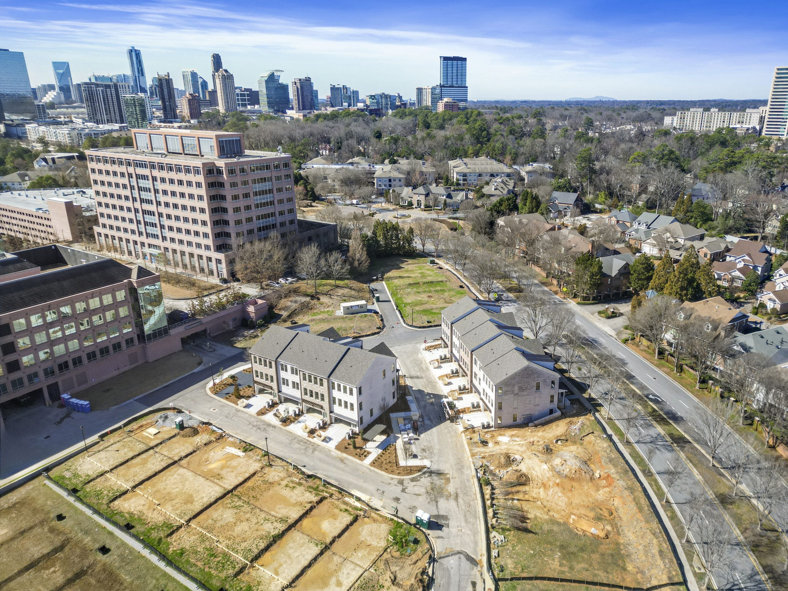 Aerial view of suburban area with buildings under construction, multiple houses in various stages of completion, surrounded by roads and empty plots, city skyline in the background with tall buildings and mixed residential and commercial structures.