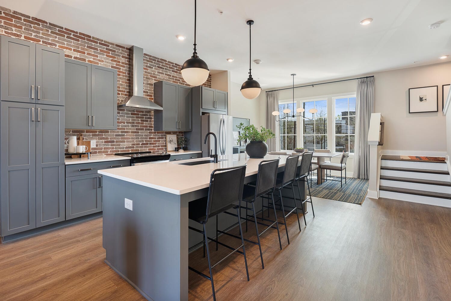 Kitchen and dining area with grey cabinets, white countertop island, black bar stools, pendant lights, stainless steel appliances, exposed brick wall, large windows, and adjacent dining table.