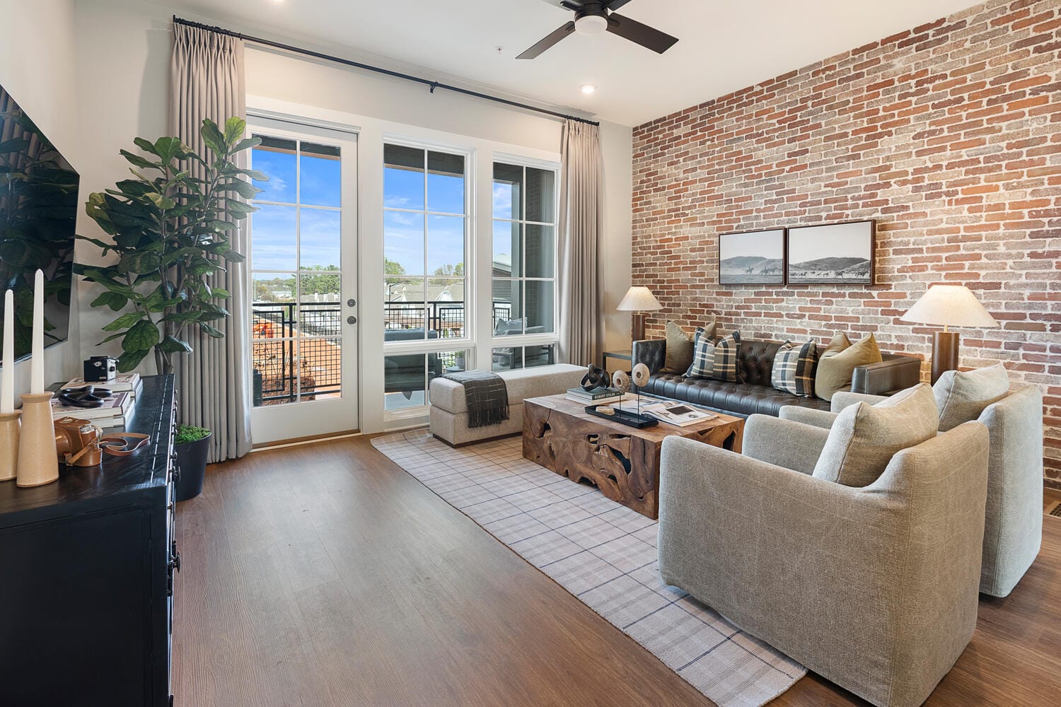 Living room with brick accent wall, large window, glass door to balcony with view, beige sofas, wooden coffee table, checkered rug, ceiling fan, plants, candles, lamps, and framed pictures