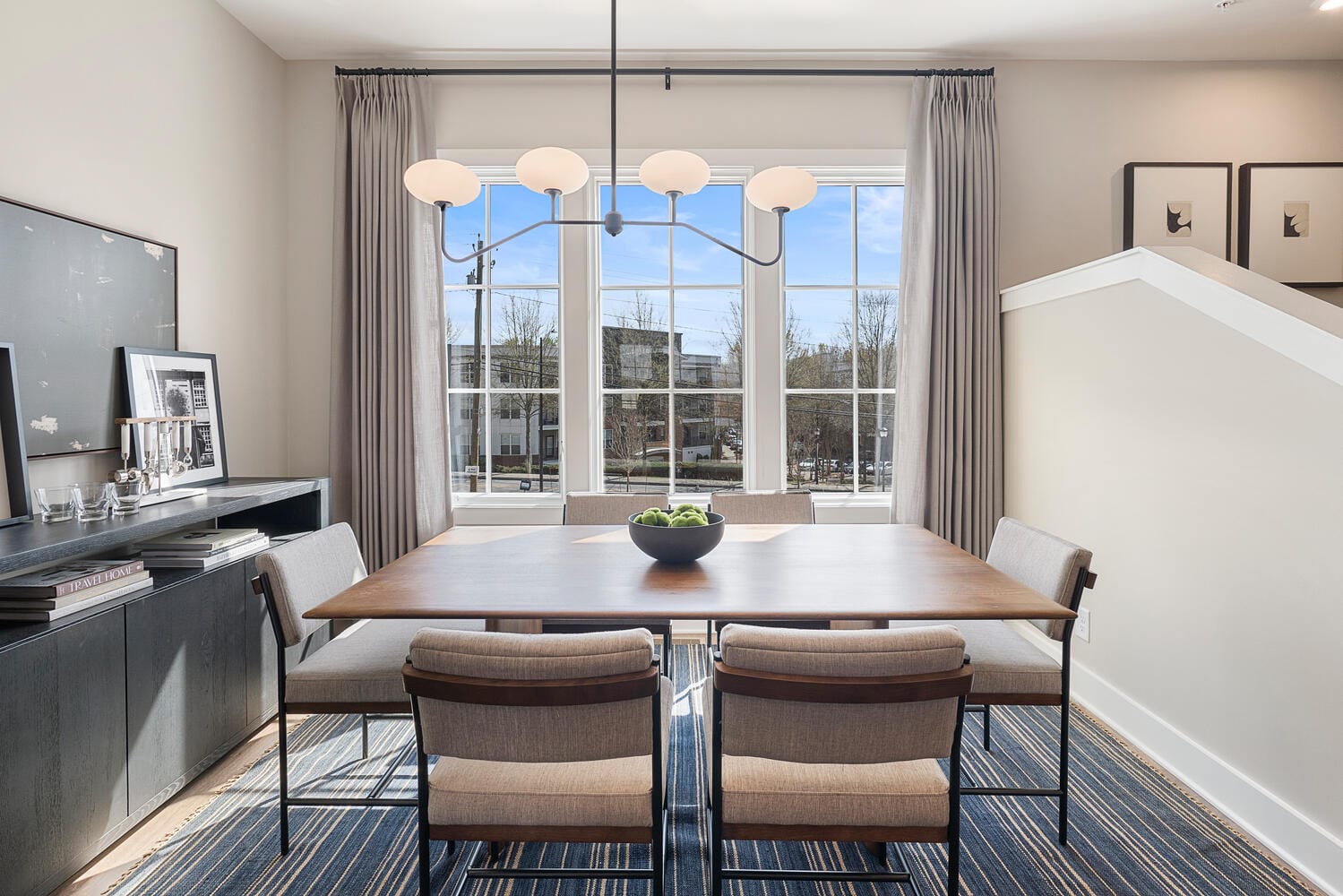 Dining room with rectangular wooden table, six chairs, bowl of green apples, large window with curtains, contemporary chandelier, sideboard with framed pictures, decorative items, and striped rug.