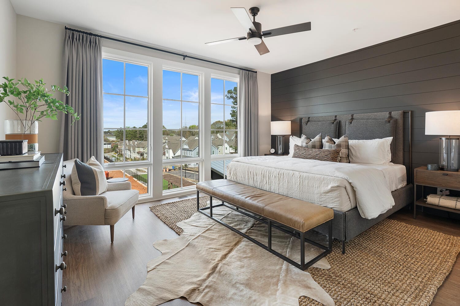 Bedroom with large bed, bench, chair, large windows with curtains, ceiling fan, and nightstands with lamps.