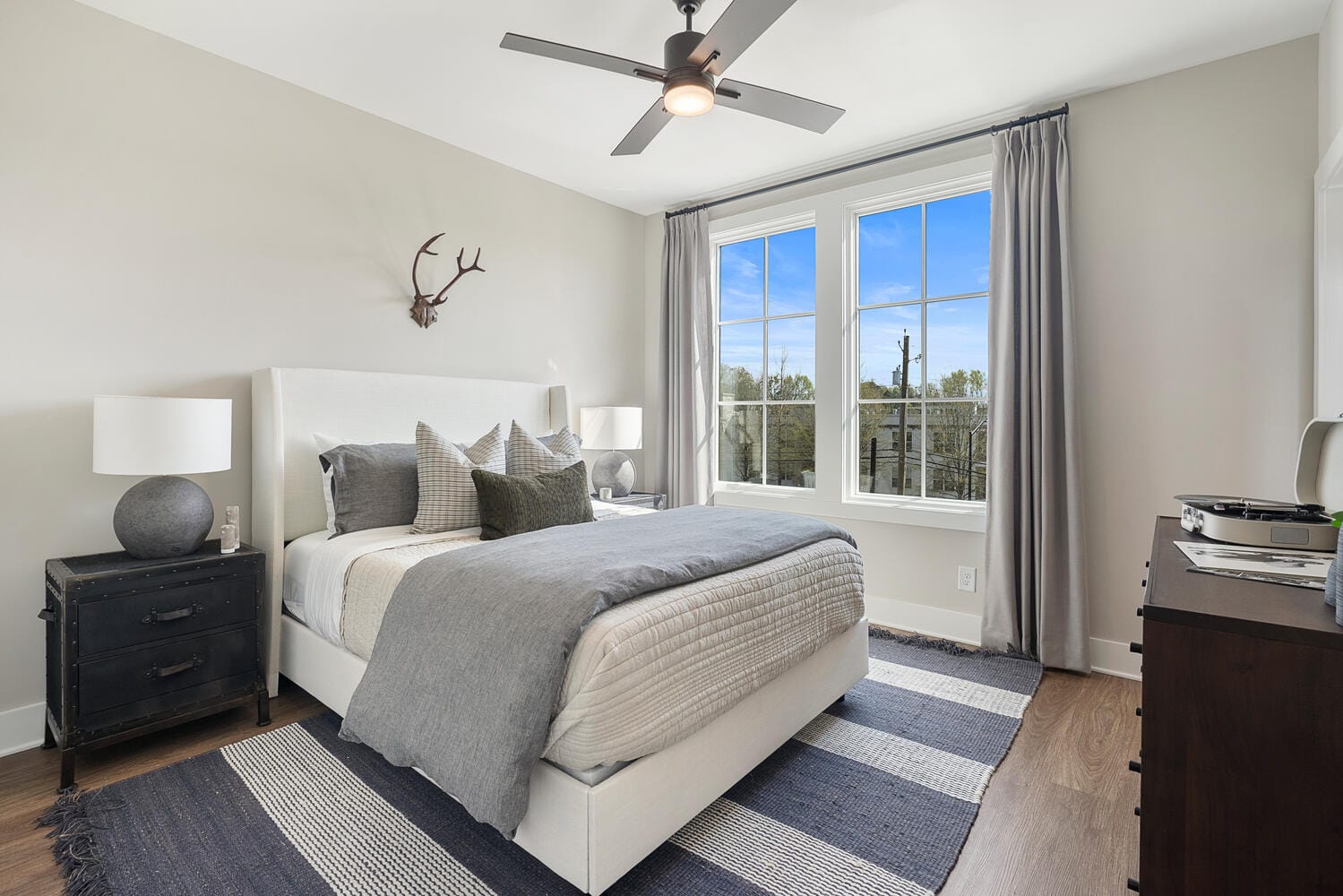 Bedroom with large window, gray and white bedding, bedside tables with lamps, ceiling fan, and dresser.