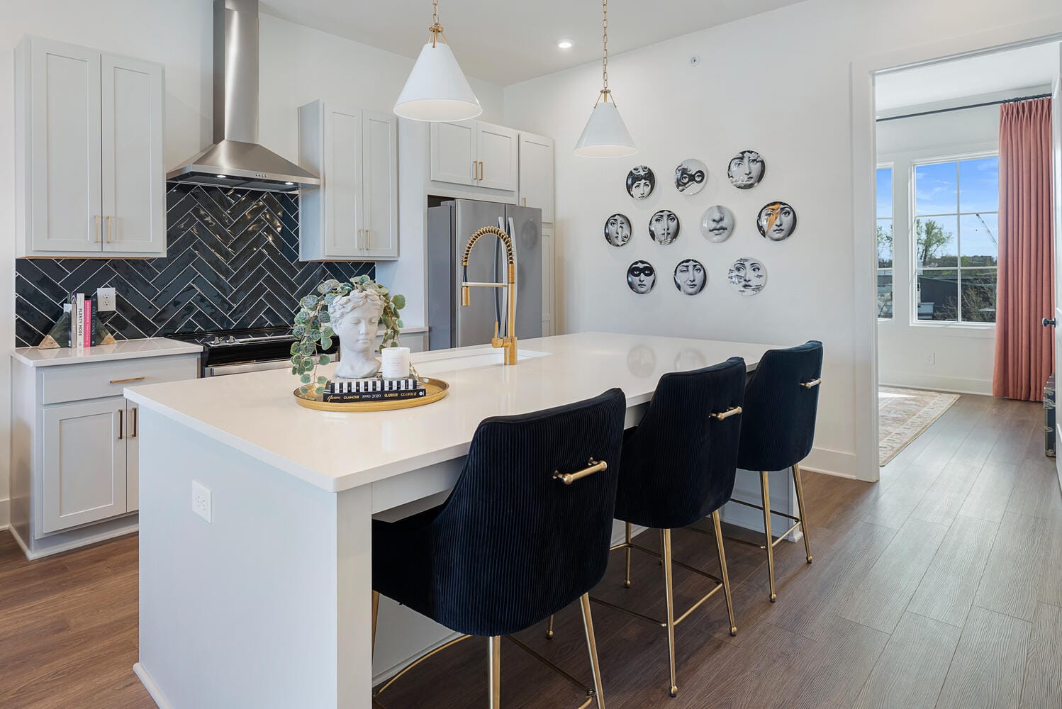 Kitchen with large island, white countertop, black chairs with gold accents, decorative tray with bust sculpture and greenery, white cabinetry, stainless steel appliances, black herringbone tile backsplash, pendant lights, artistic arrangement of round decorative plates, open doorway with pink curtains.