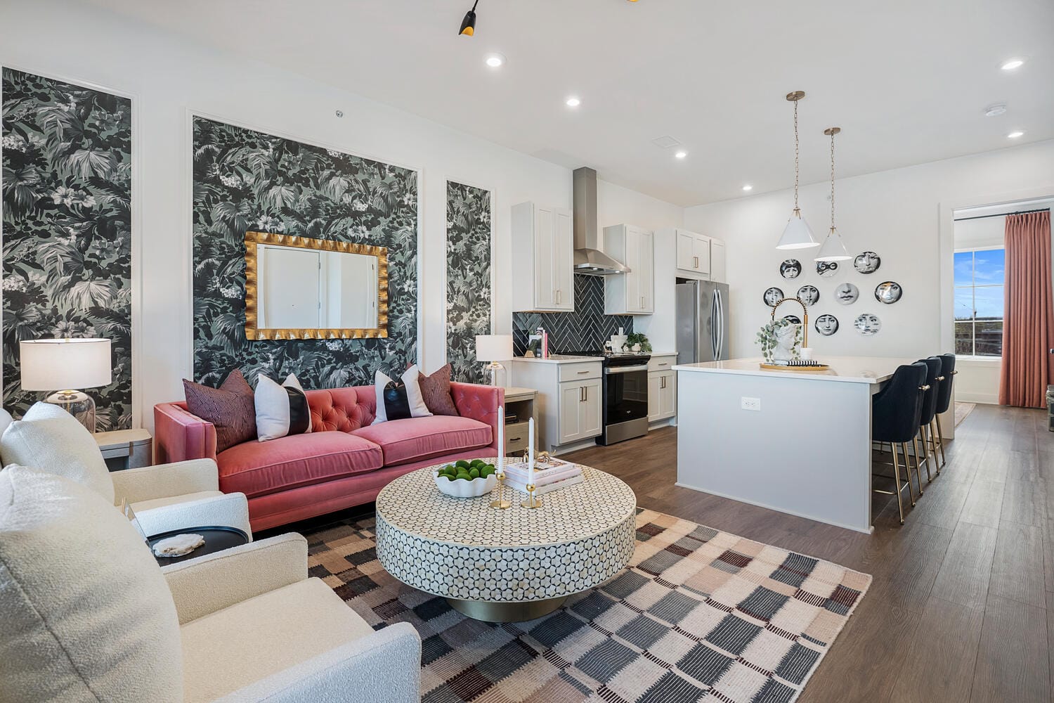 Living room and kitchen area with pink sofa, white armchair, patterned coffee table, checkered rug, floral wallpaper, large mirror, kitchen island with bar stools, pendant lights, stainless steel appliances, white cabinetry, decorative plates, and wooden flooring.