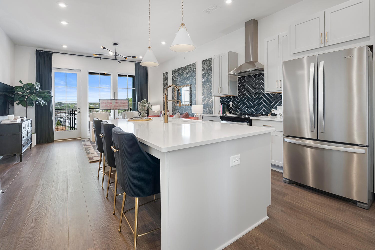Kitchen and dining area with white island, gold faucet, dark blue chairs, stainless steel appliances, herringbone tile backsplash, pendant lights, wooden floor, large windows, dining table, and decorative items.