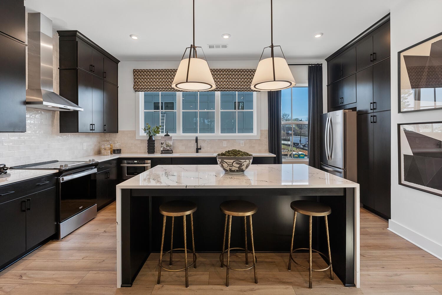 Kitchen with dark cabinetry, stainless steel appliances, large island with white countertop, wooden stools, pendant lights, white tile backsplash, windows, light wood floor, framed artworks
