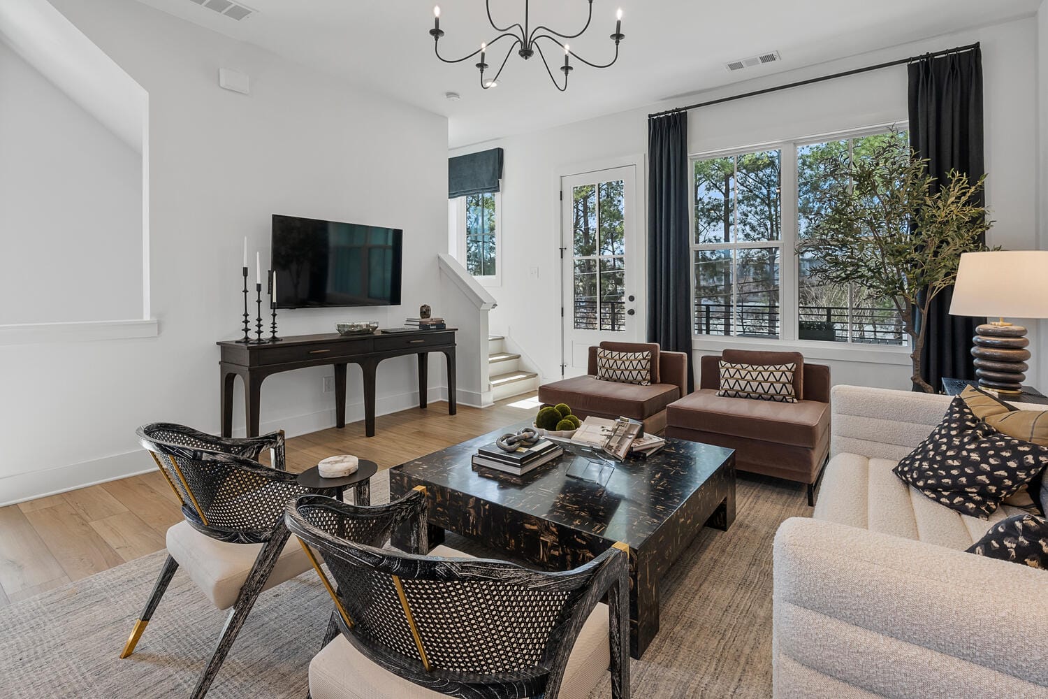 Living room with large window, dark curtains, brown chairs, beige sofa, black woven chairs with white cushions, black marble coffee table, black console table, decorative items, flat-screen TV, chandelier, wooden floor, stairs.