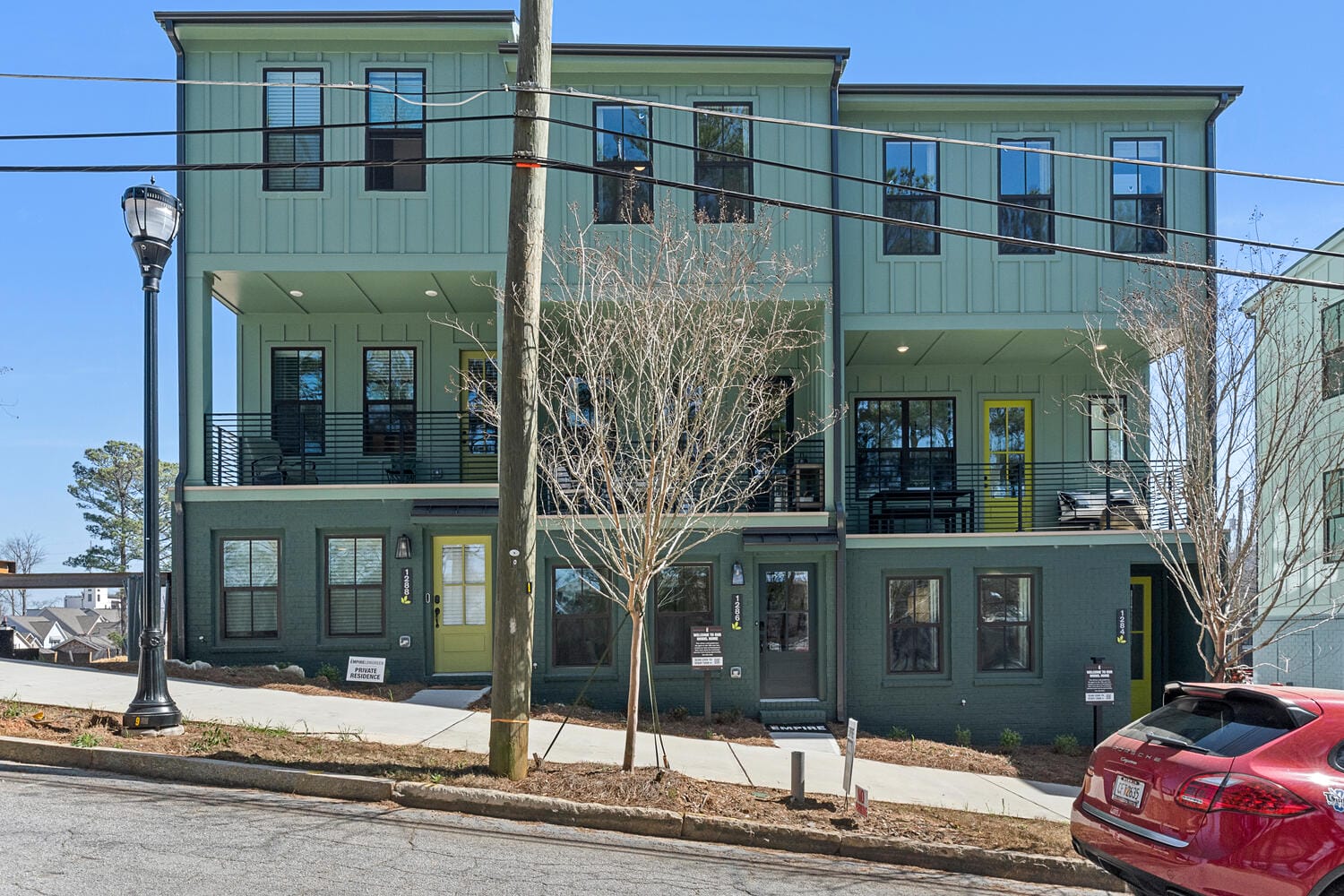Multi-story residential building with green exterior walls, large windows, balconies, sidewalk, red car, streetlamp, utility pole.