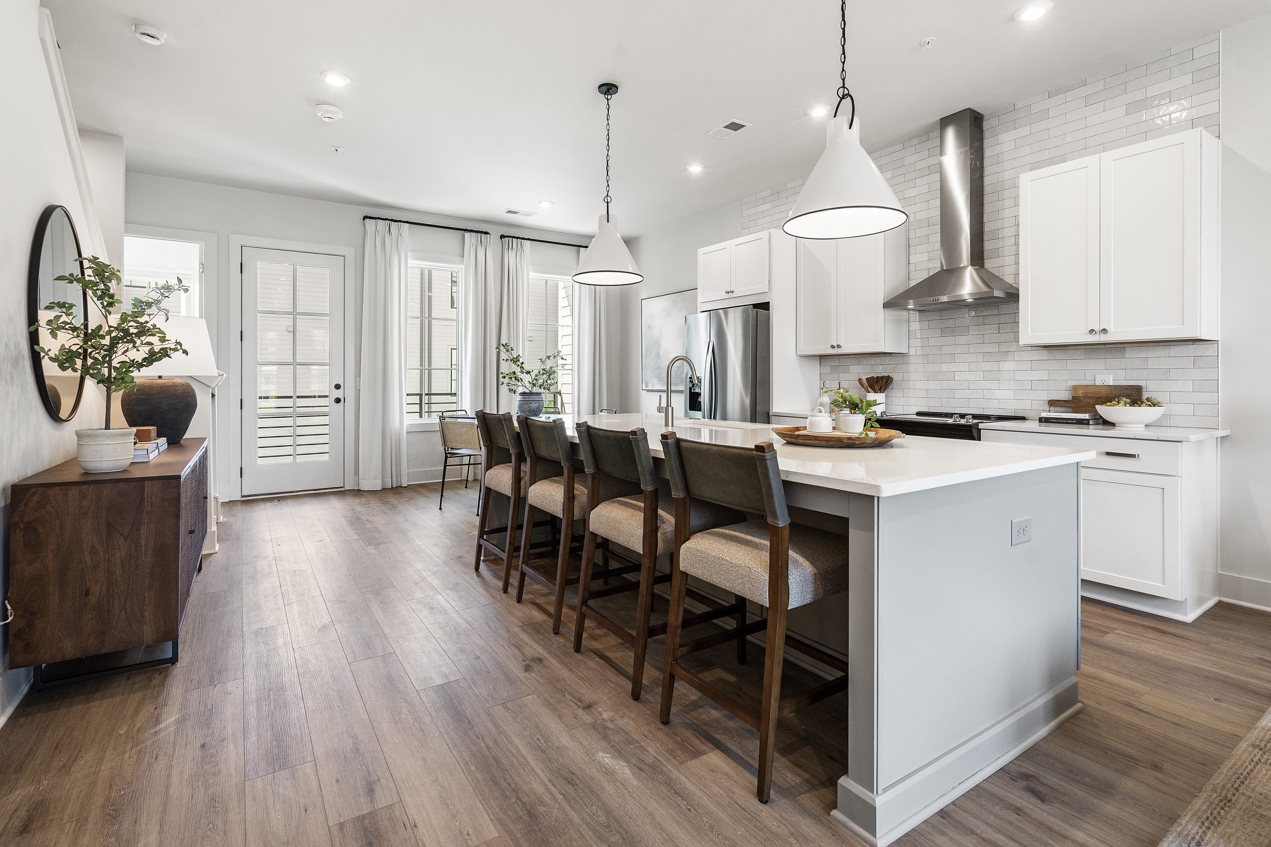 Kitchen and dining area with large island, bar stools, pendant lights, white cabinetry, stainless steel appliances, subway tile backsplash, wooden sideboard, decorative plants, round mirror, light wood floor, French doors.