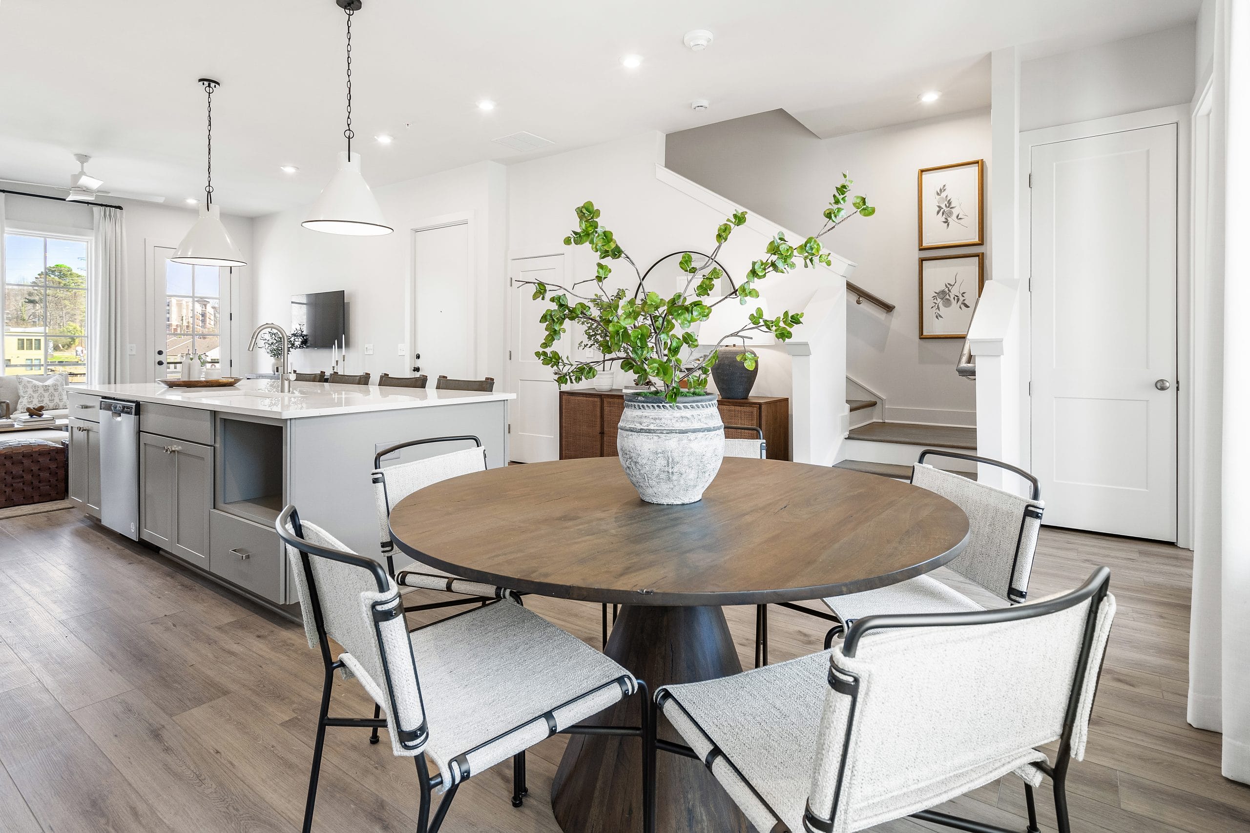 Kitchen and dining area with round wooden table, cushioned chairs, vase with green foliage, white cabinetry, large island with sink and seating, pendant lighting, living area with sofa and TV, staircase, framed artworks, and light-colored wood flooring.