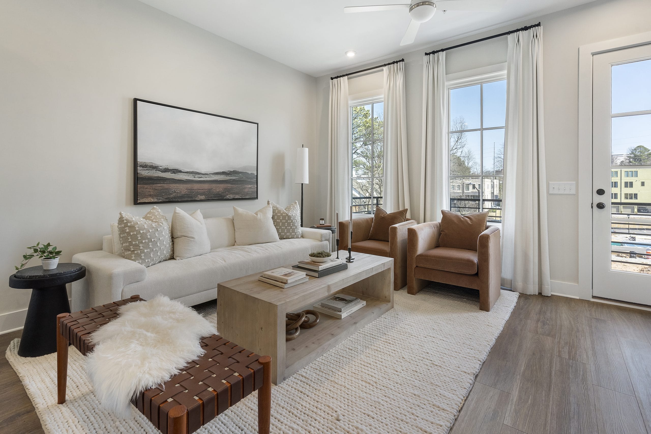 Cozy living room with a beige sofa, throw pillows, brown armchairs, wooden coffee table, woven bench with white fur accents, side table with a plant, landscape painting, big windows with white curtains, glass door, light wood floor, and a textured rug.