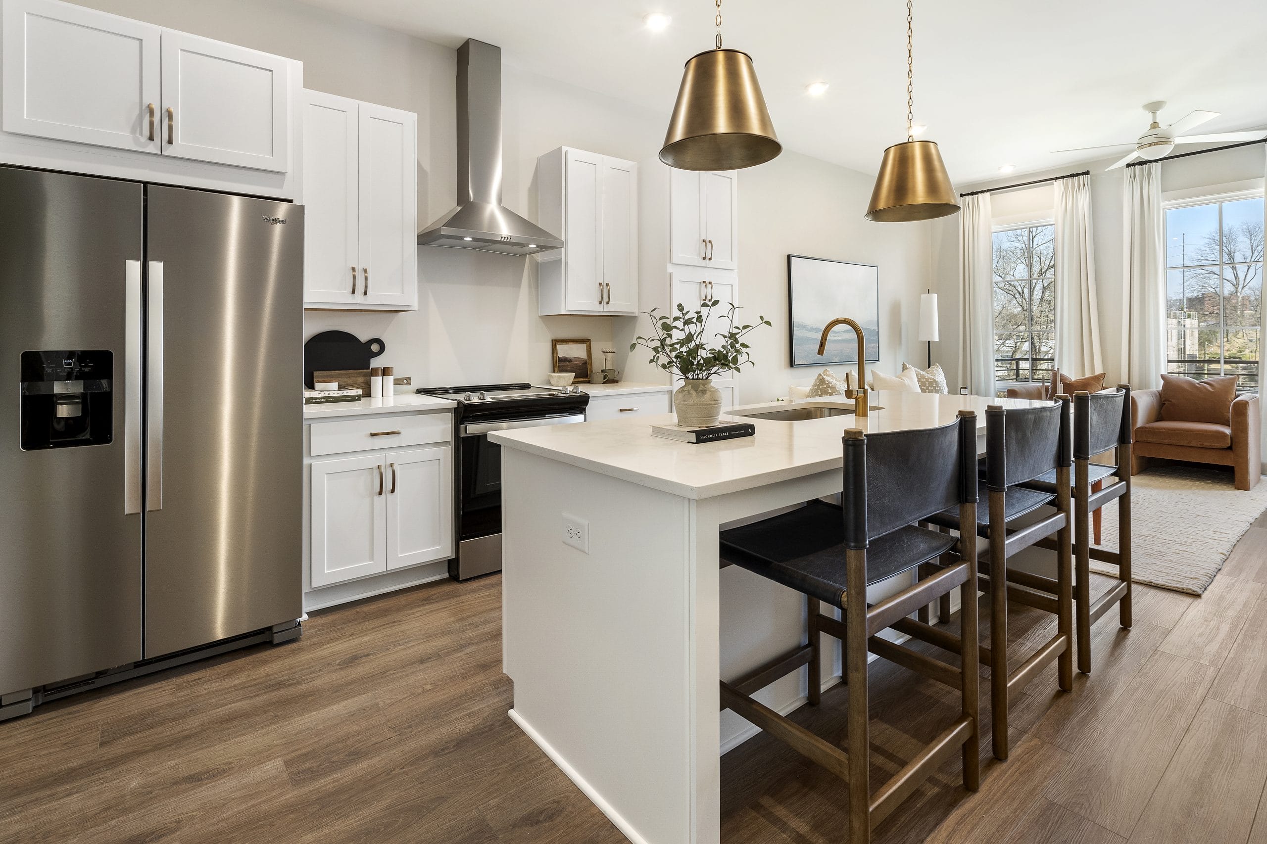 Kitchen with white cabinetry, stainless steel appliances, central island with white countertop, wooden bar stools with dark cushions, pendant lights with gold shades, range hood above stove, and open living area with large windows.