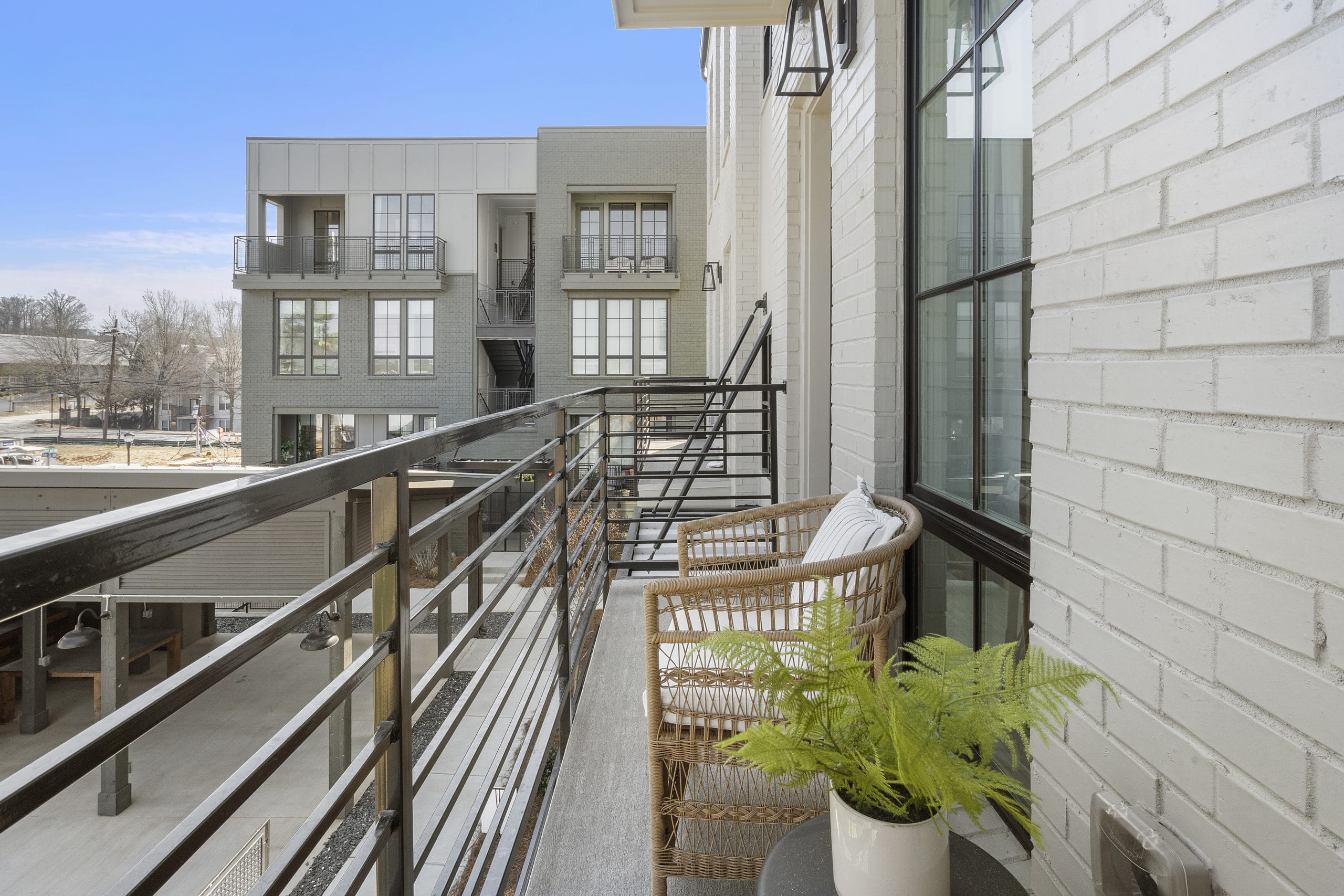 Balcony with wicker chairs, small table with potted fern, black metal railings, view of courtyard and contemporary buildings, clear blue sky.