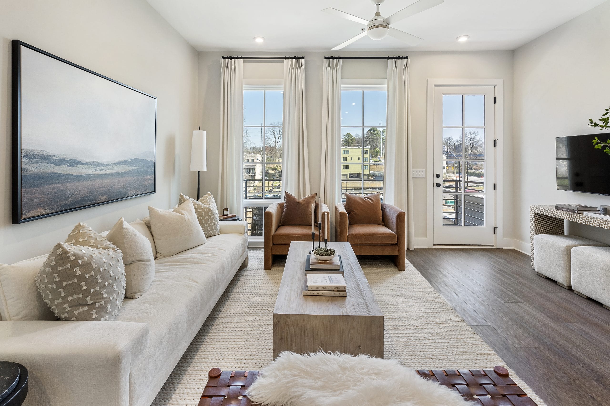 Living room with a white sofa, brown leather chairs, wooden coffee table, landscape painting, hardwood floors, light-colored rug, tall windows with white curtains, door leading outside, ceiling fan, wall-mounted TV, cushions, books, candles, and plants.