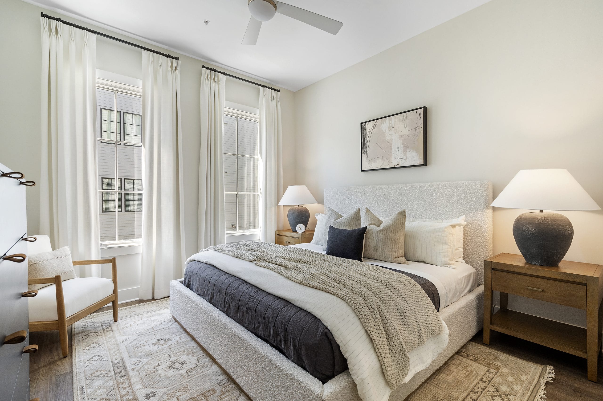 Bright bedroom with a comfy bed, white and beige bedding, pillows, wooden nightstands with lamps, chair by the window, tall windows with white curtains, ceiling fan, framed picture above the headboard, and a patterned rug.
