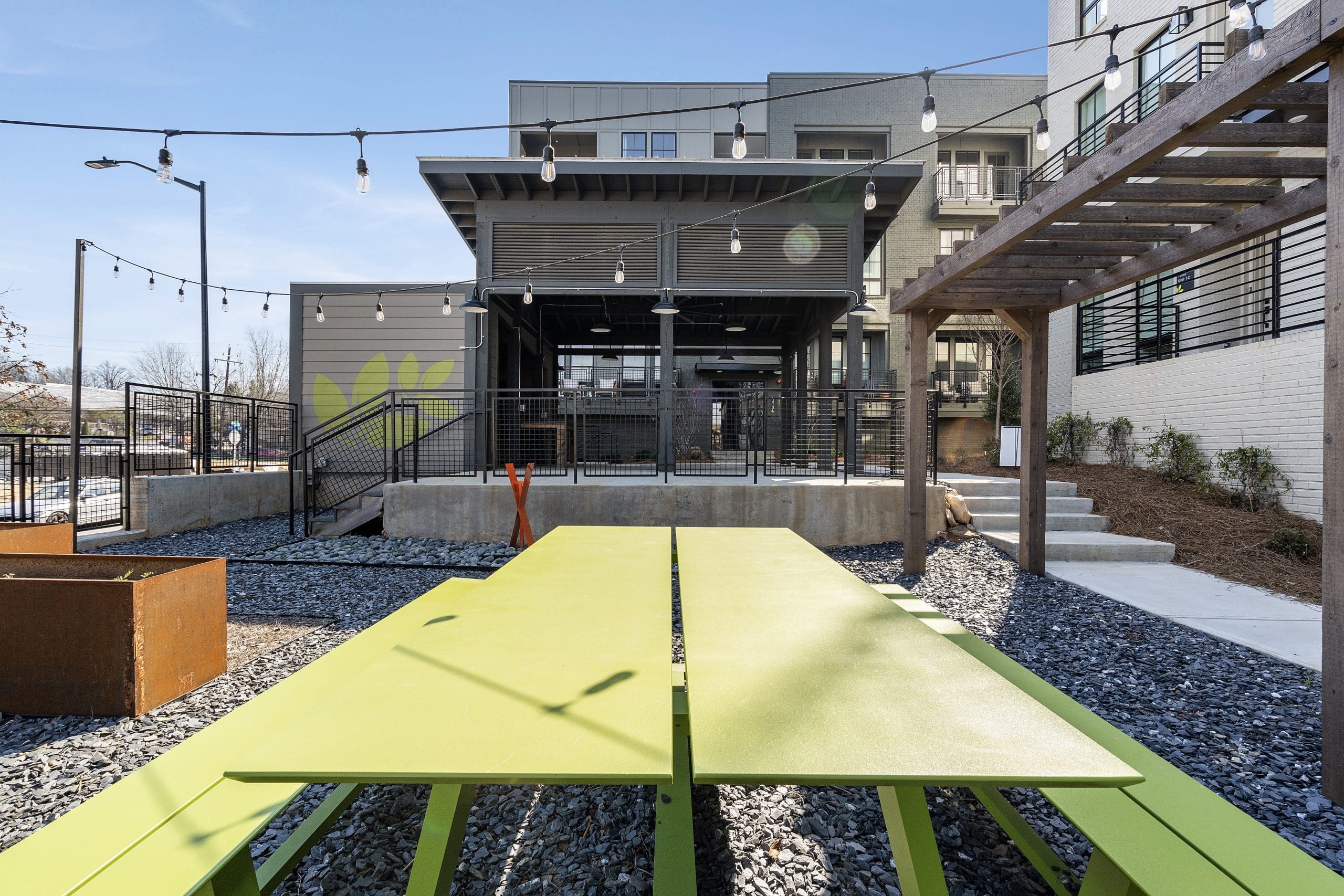 Outdoor seating area with bright green picnic tables, gravel surface, string lights, modern building with covered patio.