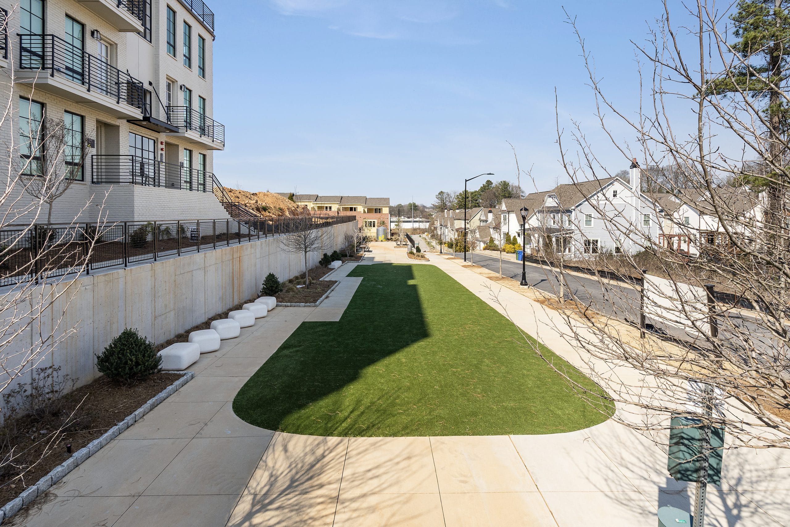 Outdoor area with green lawn, paved walkway, multi-story building with balconies, residential houses, trees, clear blue sky.