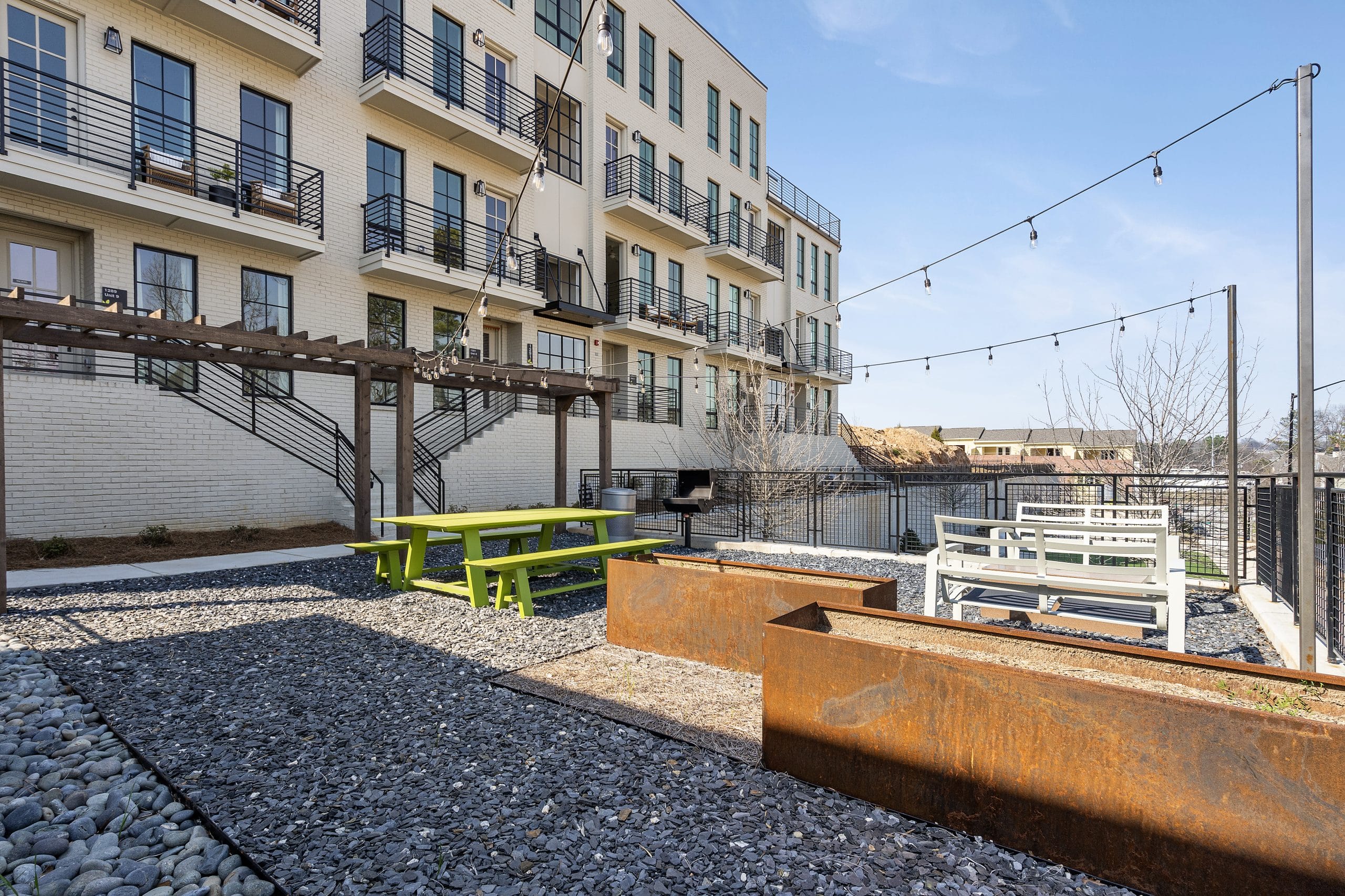 Outdoor area of modern apartment complex with balconies, green picnic table, white benches, pergola, string lights, rust-colored planters, gravel ground, small trees, view of buildings and sky.