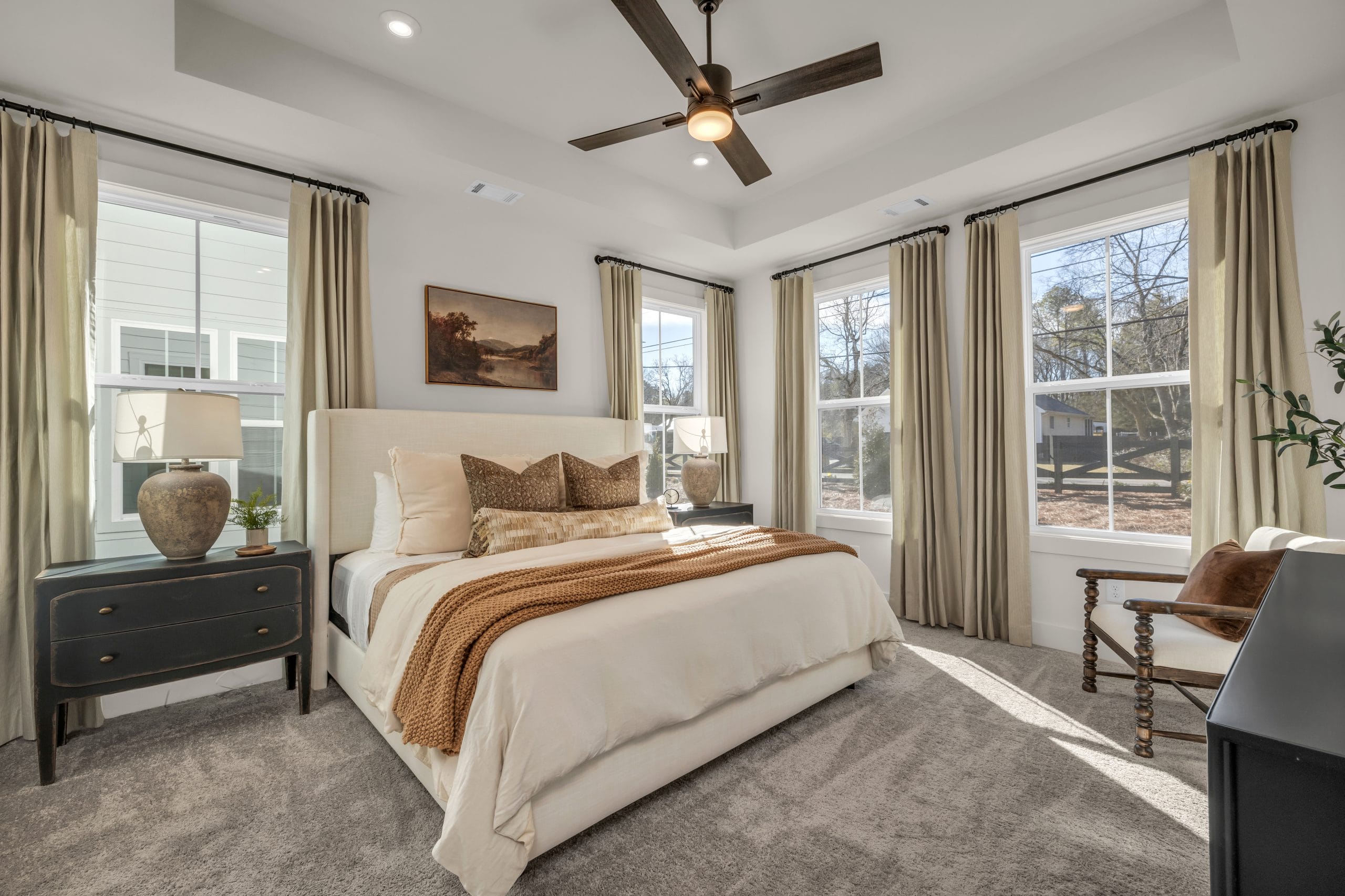Well-lit bedroom with white linens, dark nightstand, and large windows.
