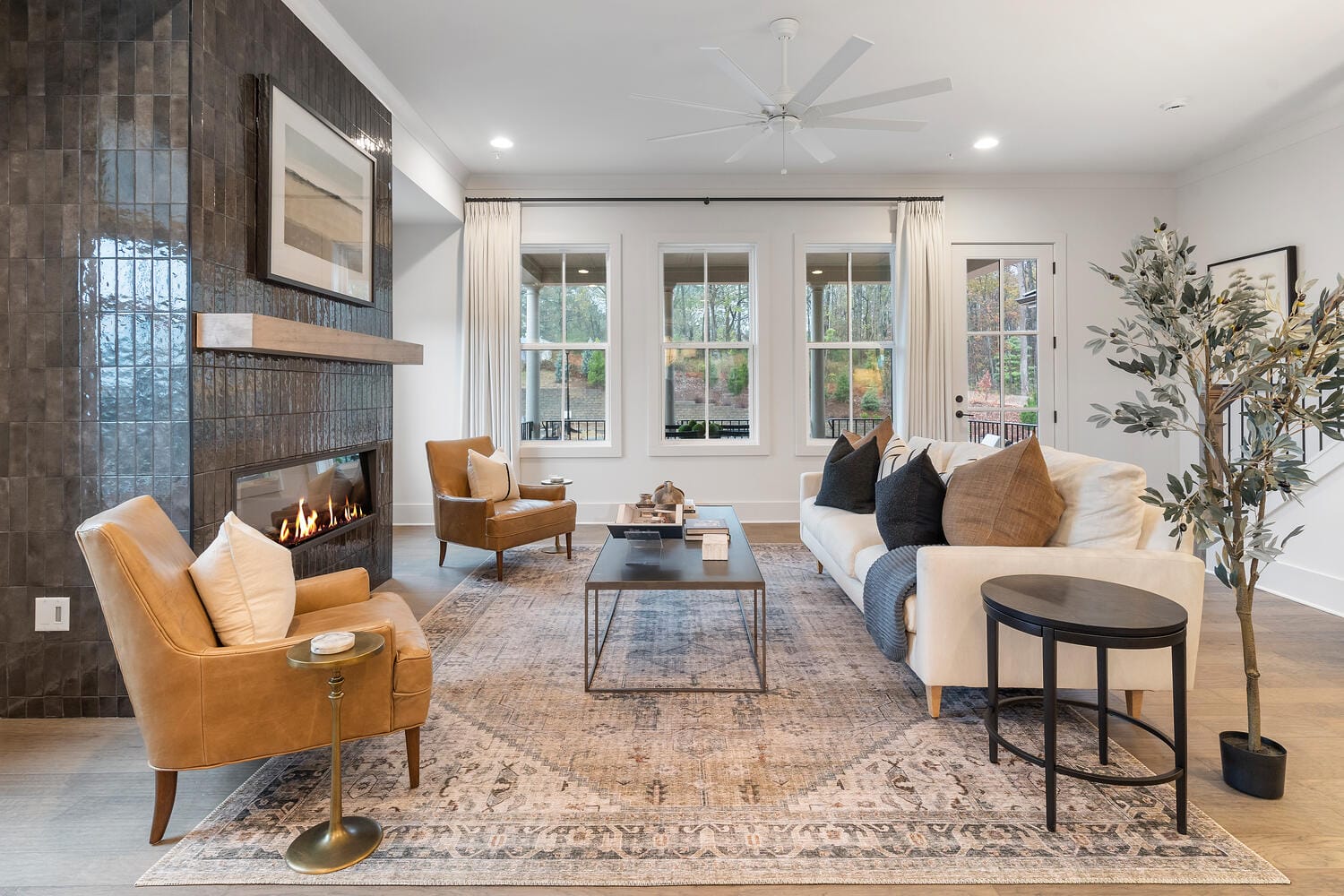 Modern living room with cozy design, fireplace with dark tiled wall, brown leather armchairs, large beige sofa with cushions, rectangular coffee table on patterned rug, round side table, artificial tree, large windows with white curtains, recessed lighting, and ceiling fan.