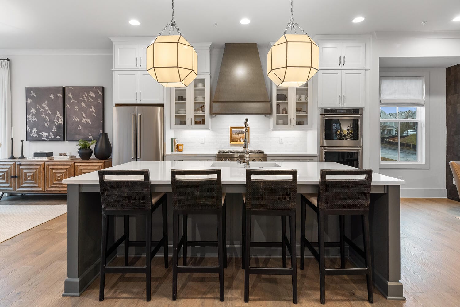 Kitchen with large island, dark-colored chairs, geometric pendant lights, white cabinetry, stainless steel appliances, and window.