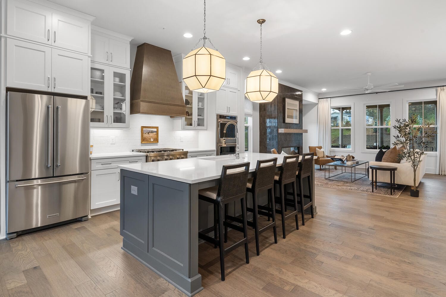 Modern kitchen with white cabinets, stainless steel fridge, island with bar stools, and living area with sofas and large windows.