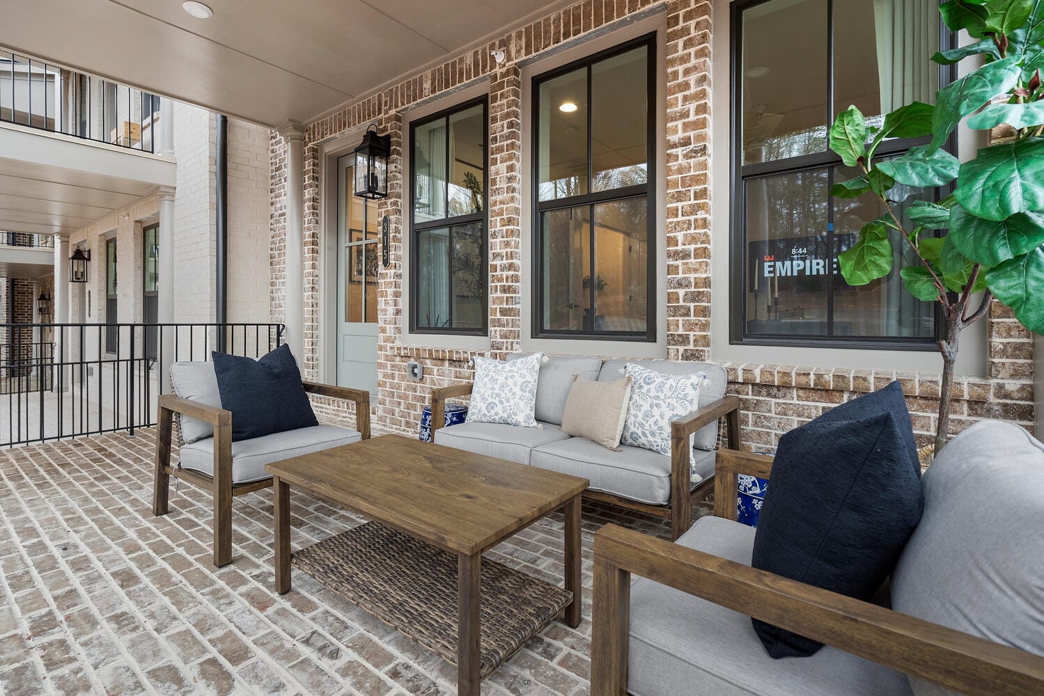 Outdoor patio with brick flooring, wooden furniture with gray cushions, coffee table, large windows, and potted plant.