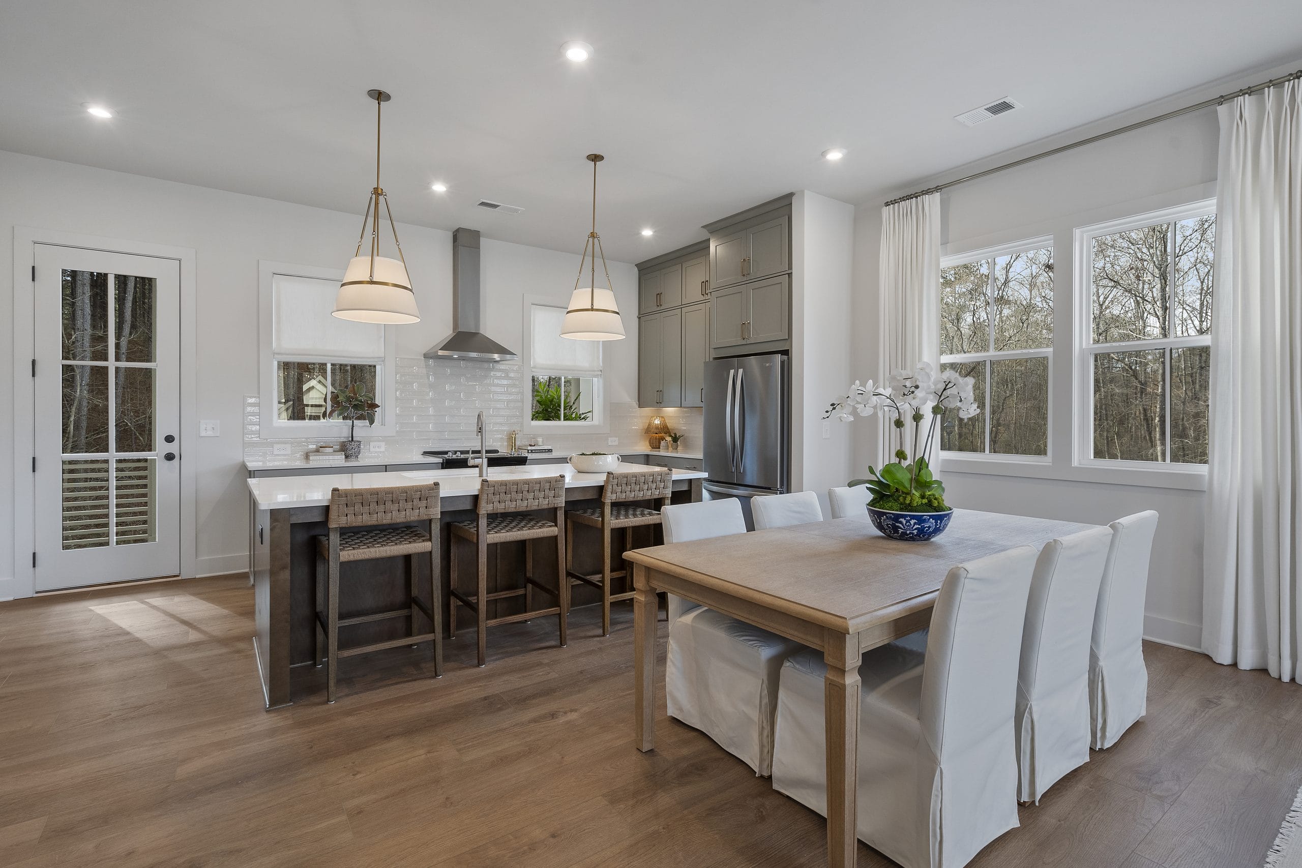 Kitchen and dining area with island, woven bar stools, pendant lights, stainless steel appliances, white cabinetry, wooden table, slipcovered chairs, large windows, and glass door.