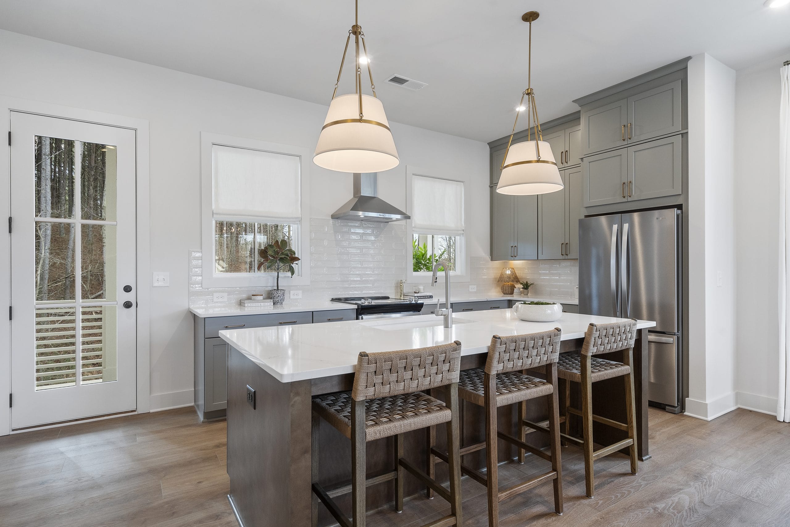Kitchen with large island, woven bar stools, pendant lights, gray cabinetry, stainless steel appliances, subway tile backsplash, windows, and light wood flooring.