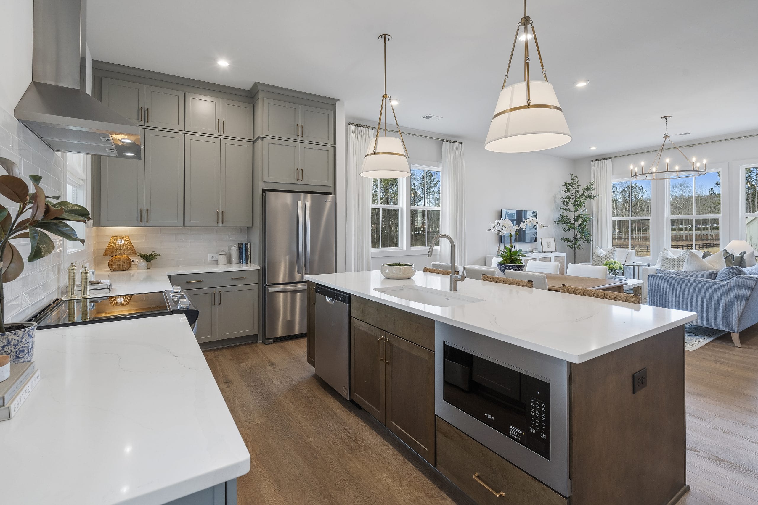 Kitchen with open layout, gray cabinetry, stainless steel appliances, large island, pendant lights, and living area with large windows and sofas.