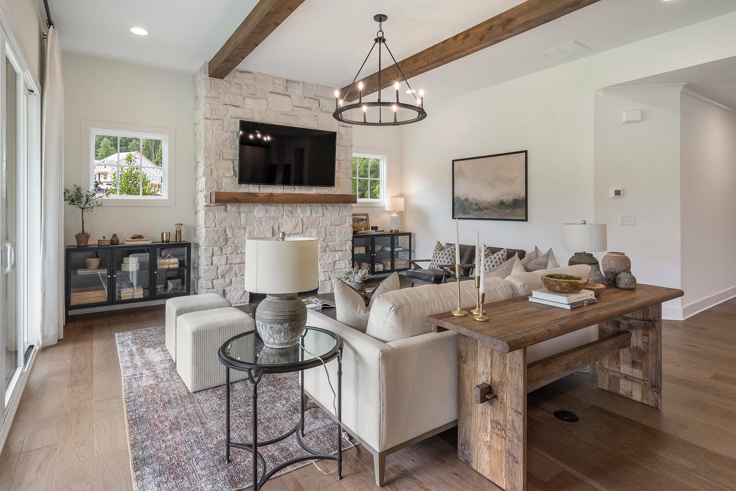 Modern living room with stone fireplace, mounted TV, beige sectional sofa, patterned throw pillows, wooden coffee table, side tables, decorative items, large windows, exposed wooden beams, rug, hardwood floor, artwork.