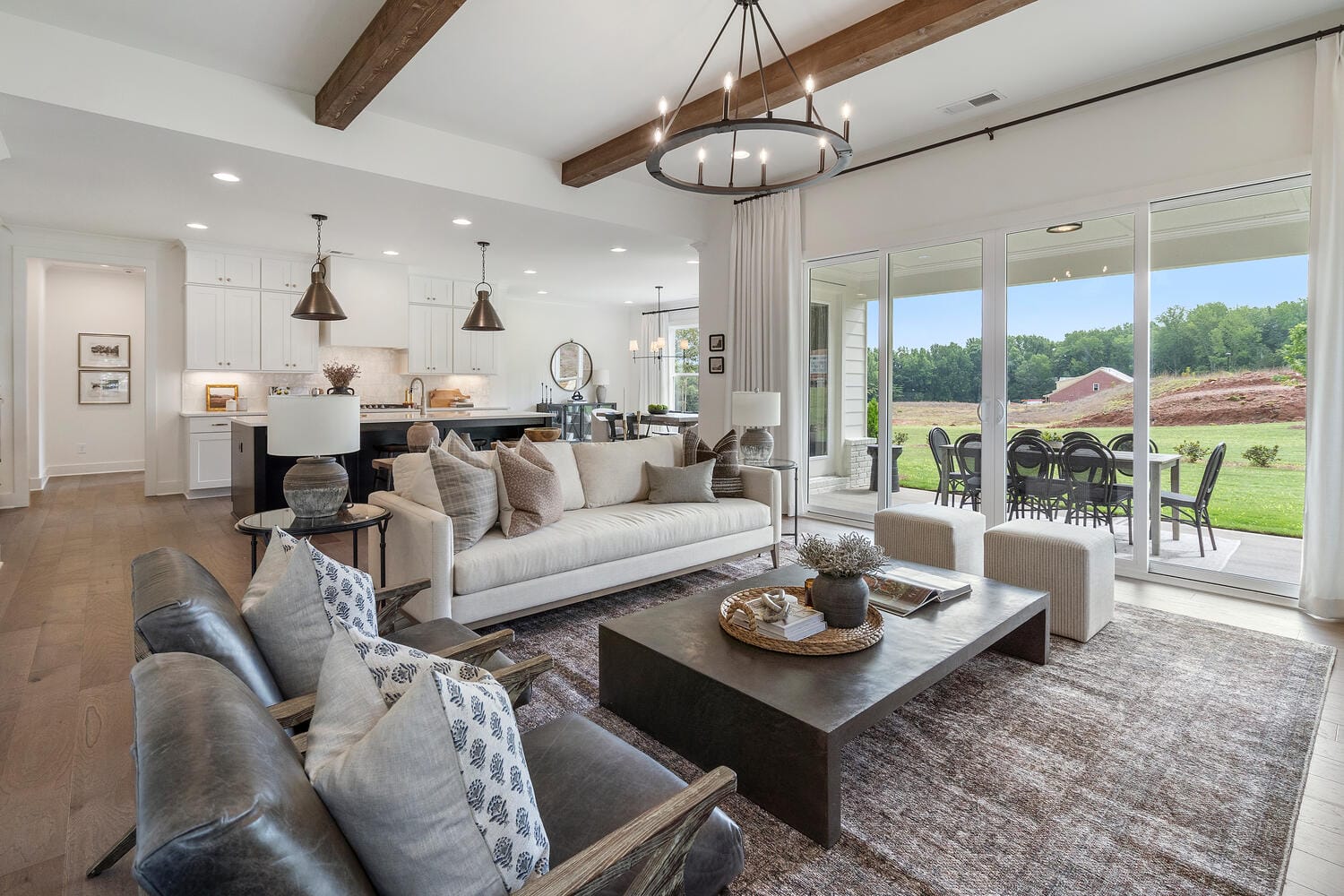 Spacious living room opening into kitchen and dining area, featuring sectional sofa, armchairs, wooden coffee table, decorative items, exposed wooden beams, white cabinetry, pendant lights, island with bar stools, stainless steel appliances, sliding glass doors to outdoor patio.