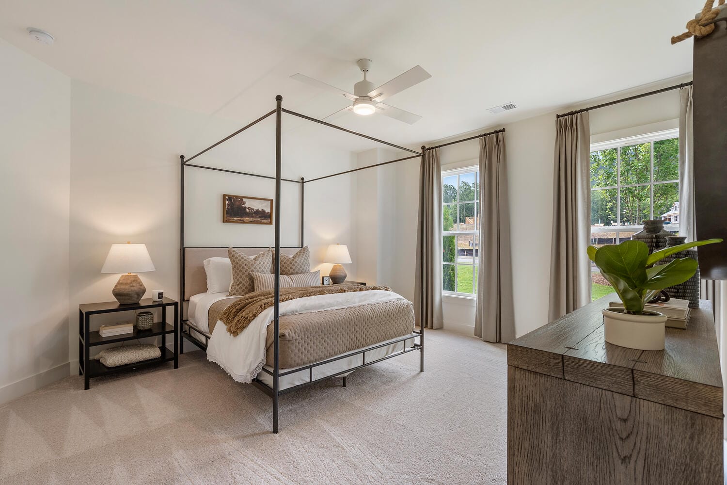 Well-lit bedroom with four-poster bed, white and beige bedding, bedside tables with lamps, ceiling fan, large windows with curtains, wooden dresser with potted plant, and light-colored carpet.