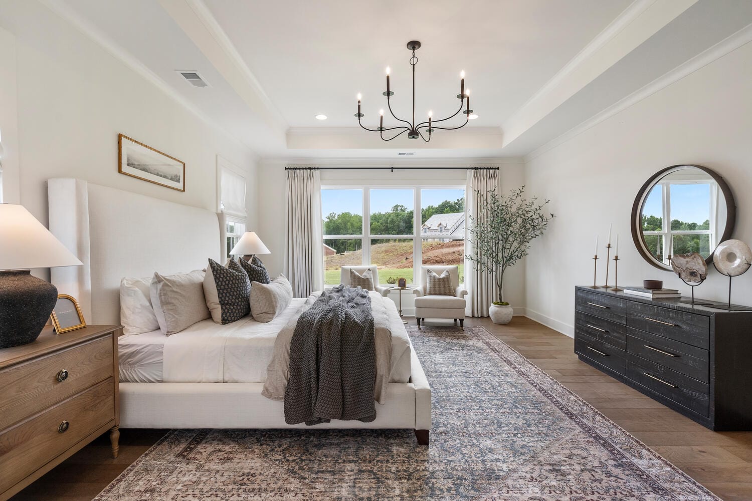 Well-decorated bedroom with large bed, white bedding, bedside tables with lamps, large window providing natural light, dark-colored dresser with decorative items, wooden floor, area rug, and chandelier.