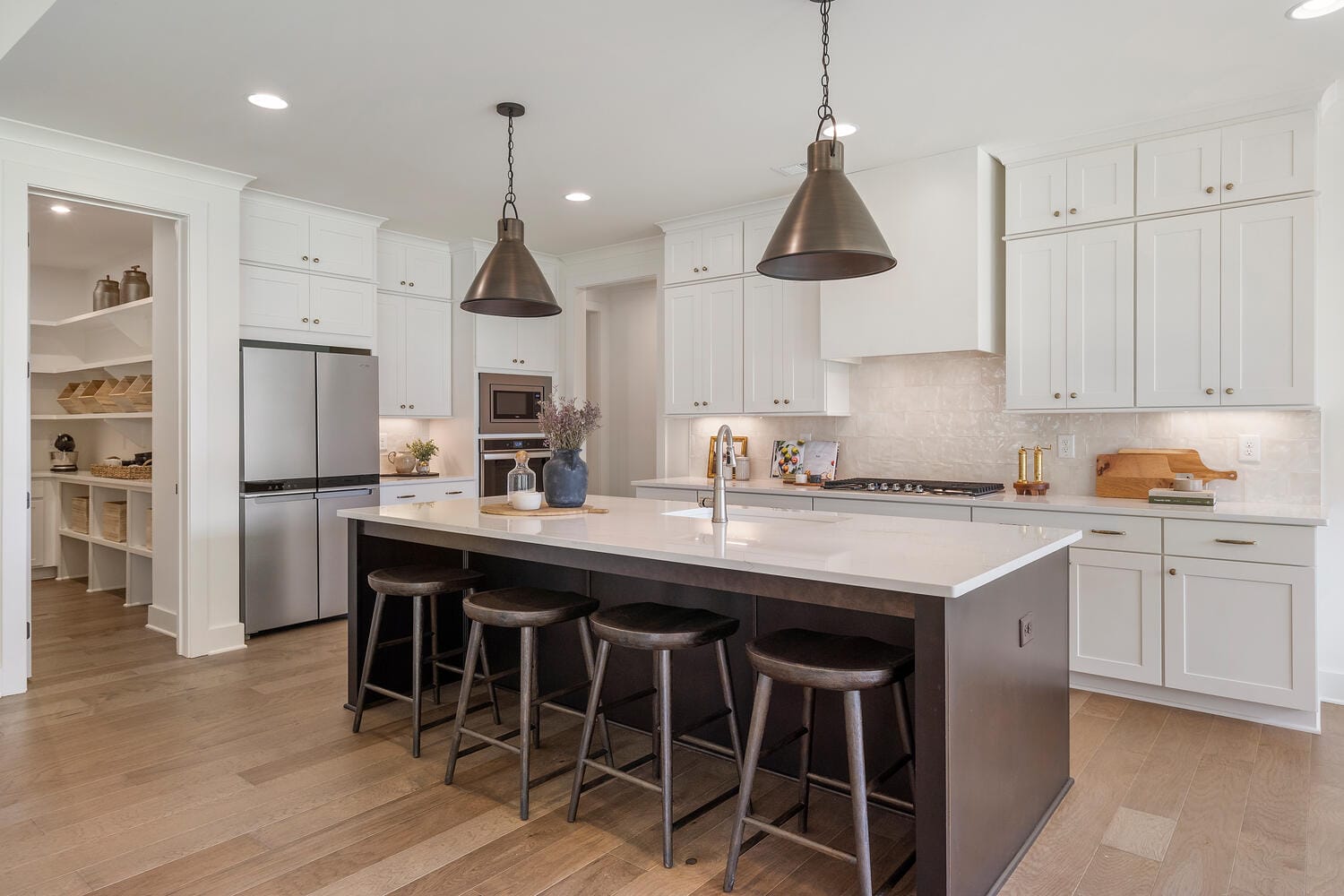 Modern kitchen with white cabinetry, stainless steel appliances, large island with white countertop, black bar stools, built-in sink, pendant lights, open pantry, wooden flooring, recessed lighting.