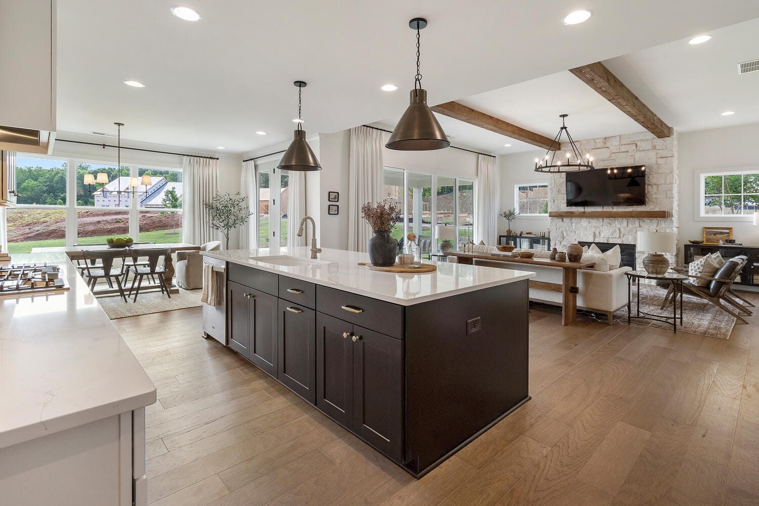 Modern open-concept kitchen and living area with large island, white countertop, dark cabinetry, pendant lights, stone fireplace with mounted TV, comfortable seating, large windows, sliding glass doors, dining table, wooden flooring, and exposed wooden beams.