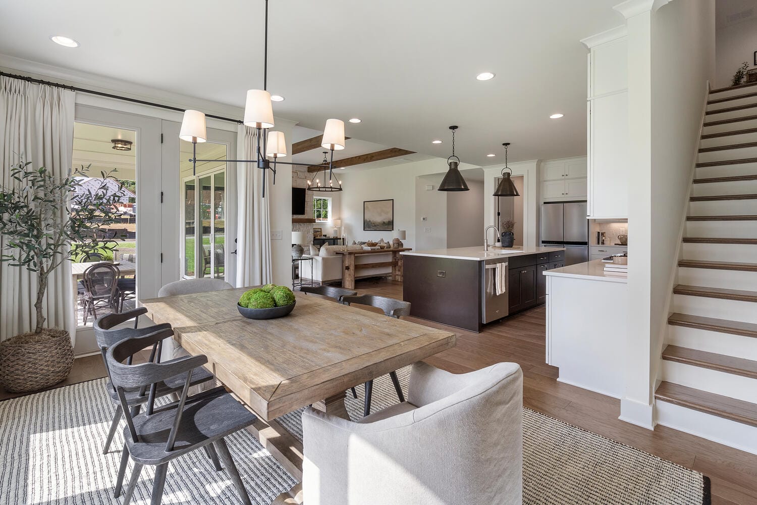Modern open-concept kitchen and dining area with wooden table, black chairs, chandelier, large glass doors to outdoor patio, kitchen island with bar stools, stainless steel appliances, white cabinetry, and staircase to upper floor.