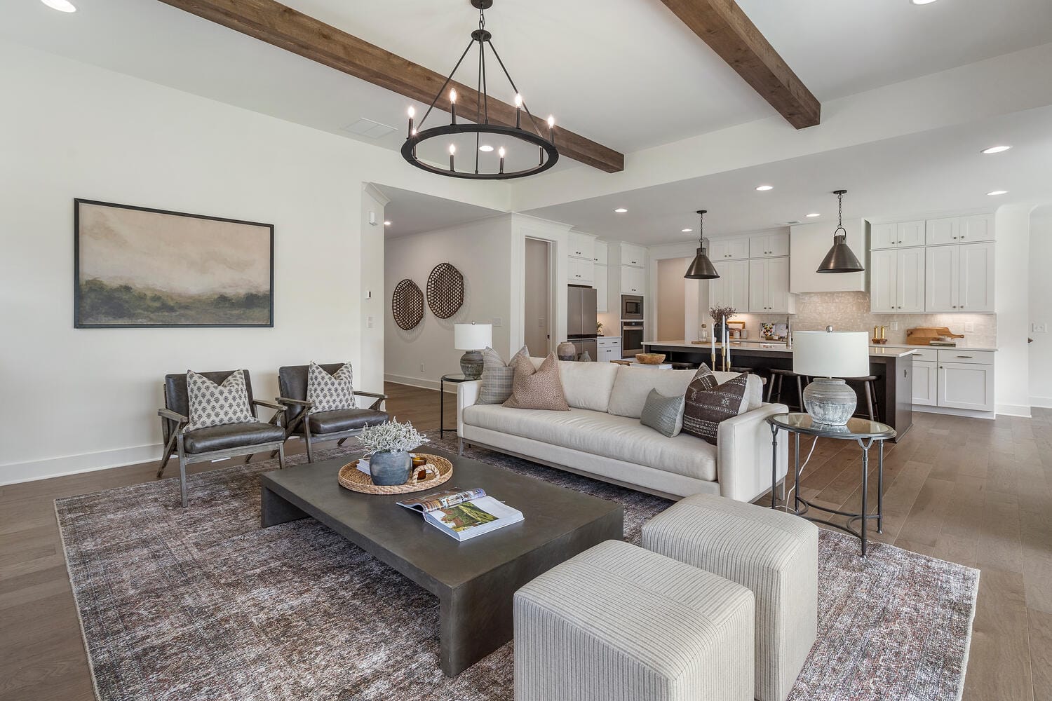 Modern open-concept living room and kitchen area featuring a large beige sofa, patterned armchairs, dark coffee table, light-colored ottomans, area rug, exposed wooden beams, chandelier, landscape painting, white cabinetry, stainless steel appliances, and kitchen island with bar stools.