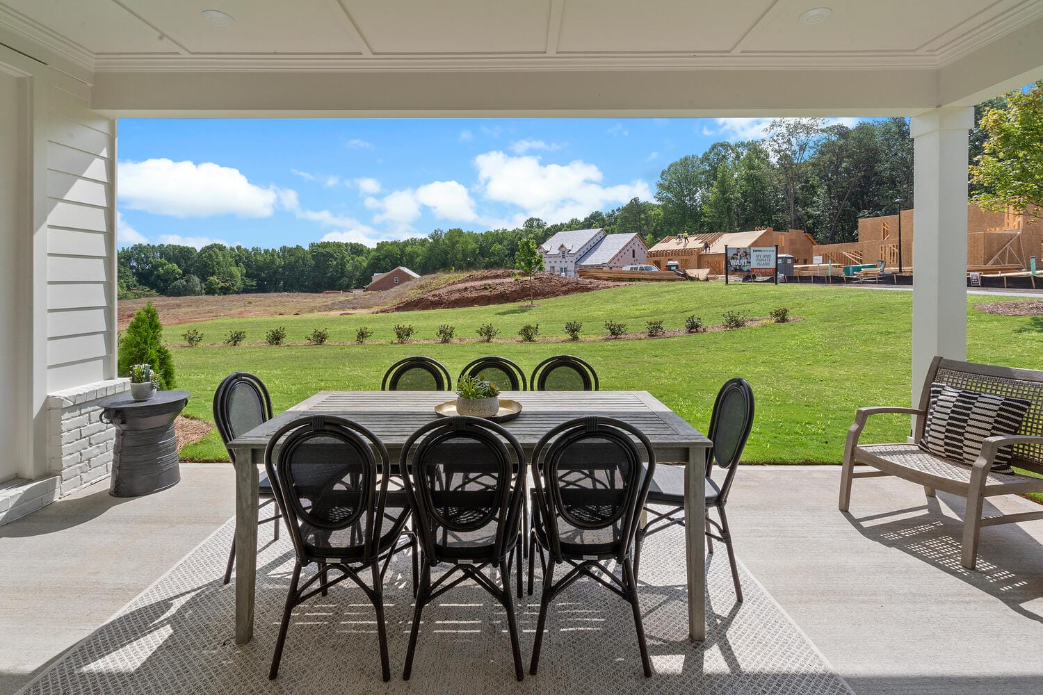 Outdoor patio area with rectangular dining table and eight black chairs, overlooking grassy yard with construction work and houses being built, bench on the right, small round table with plant, and blue sky with clouds.