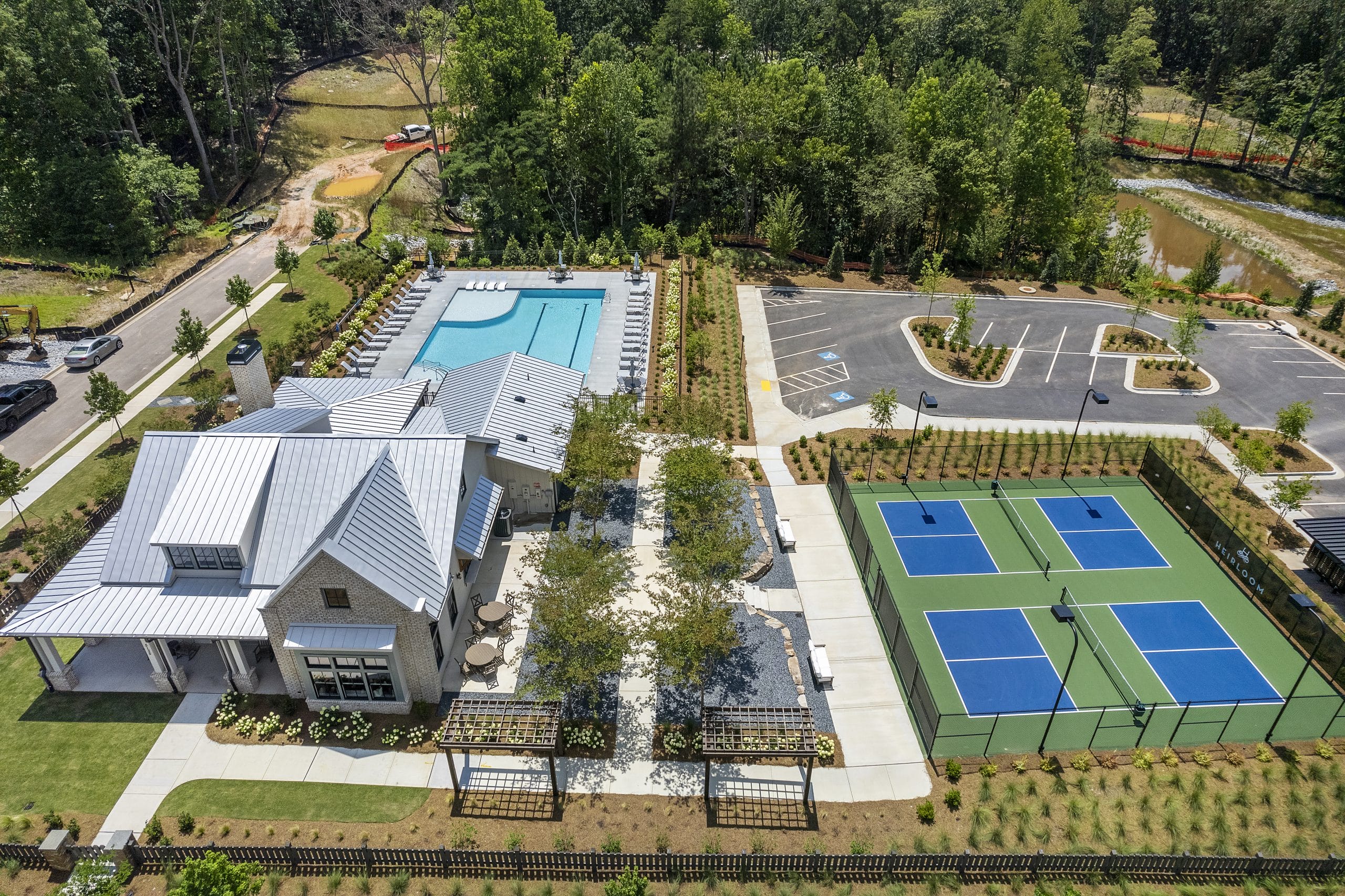 Aerial view of recreational facility with swimming pool, tennis courts, building with outdoor seating, landscaped areas, parking spaces, surrounded by trees and greenery.