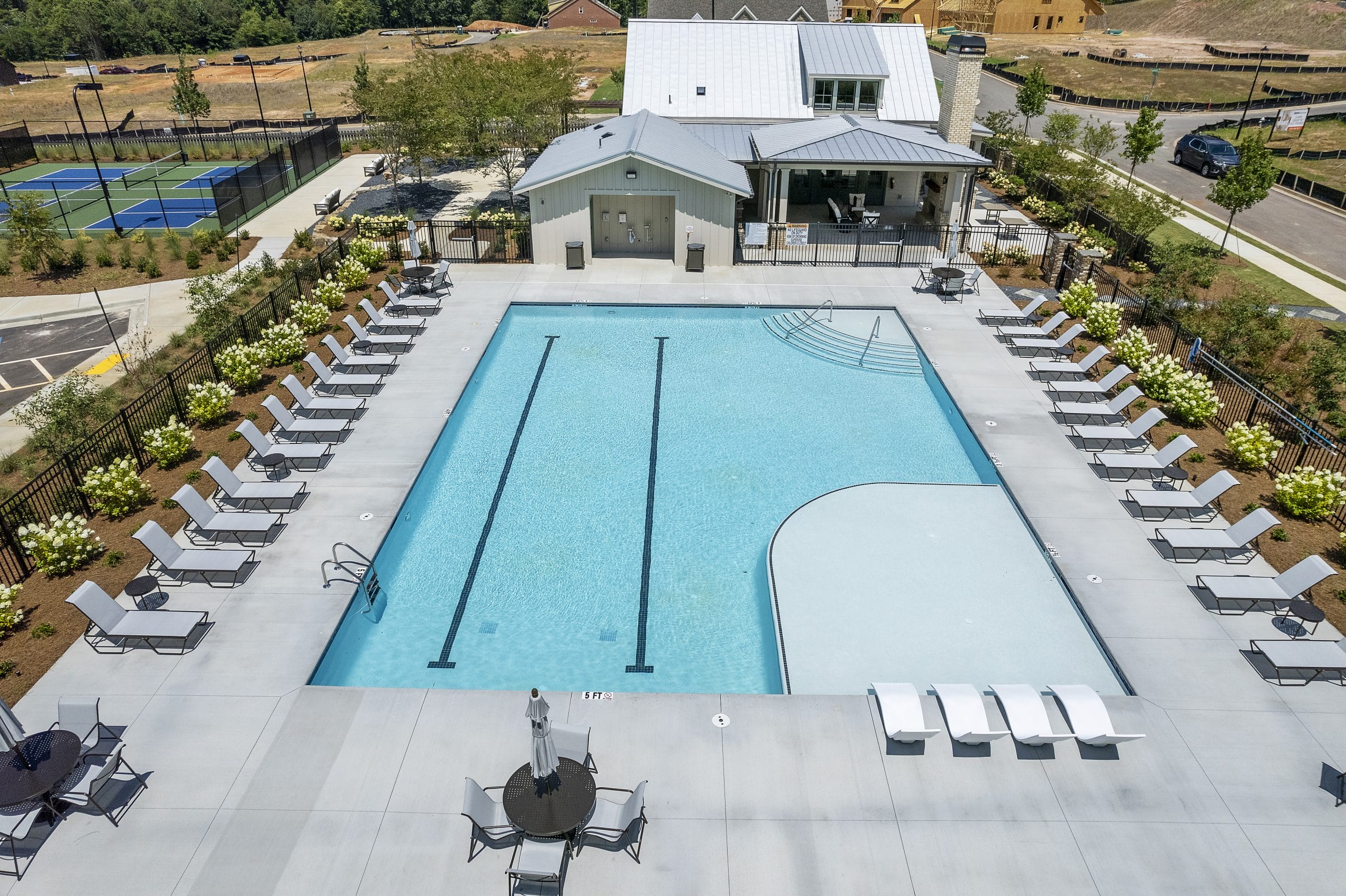 Outdoor swimming pool area with lounge chairs, tables with umbrellas, and a building with a metal roof, featuring swim lanes, shallow entry, and nearby tennis court, showcasing amenities of Empire Homes community.