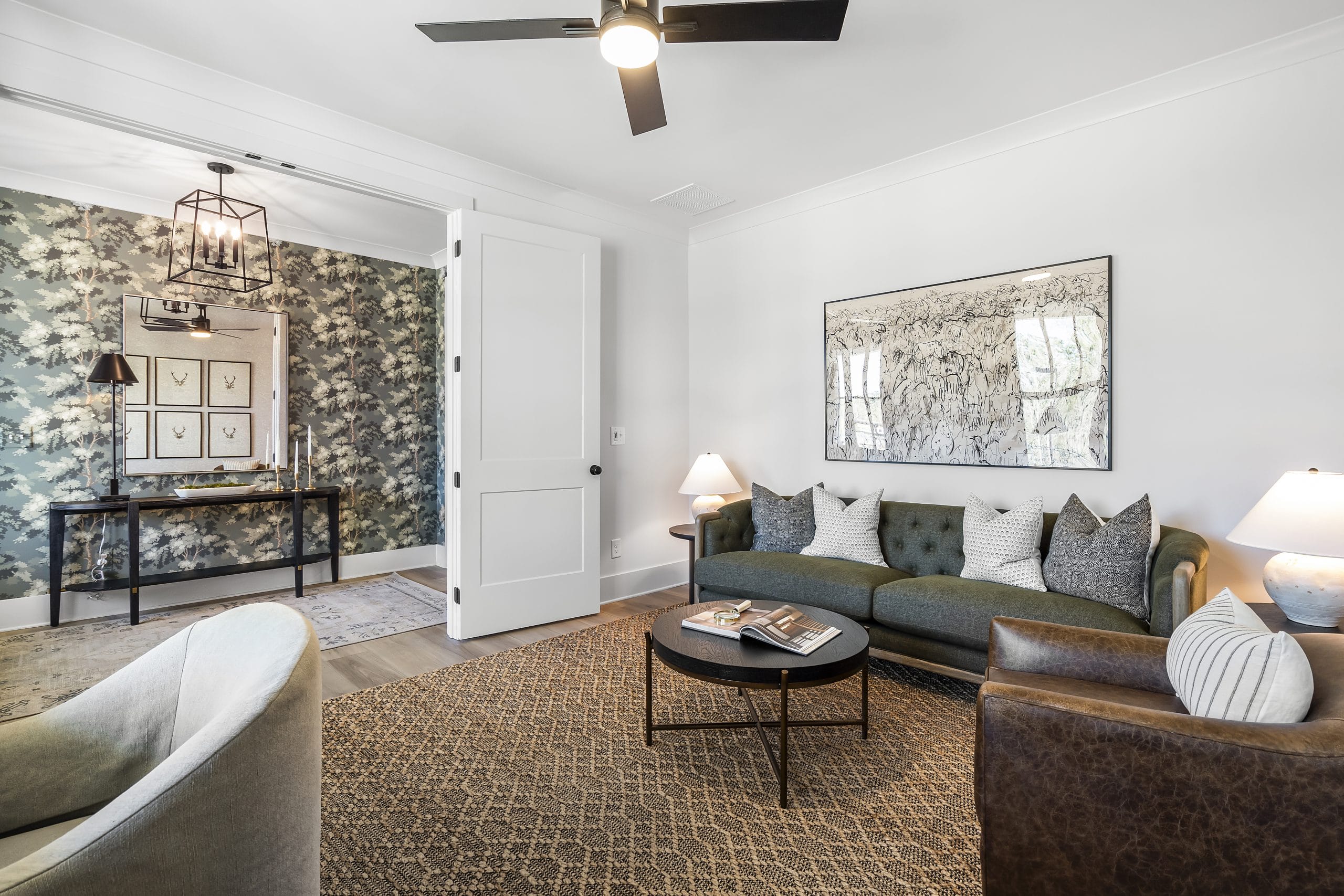 Modern living room with green sofa, round black coffee table, decorative items, white walls, ceiling fan with lights, patterned rug, adjacent area with nature motif wallpaper, console table, and framed artwork.