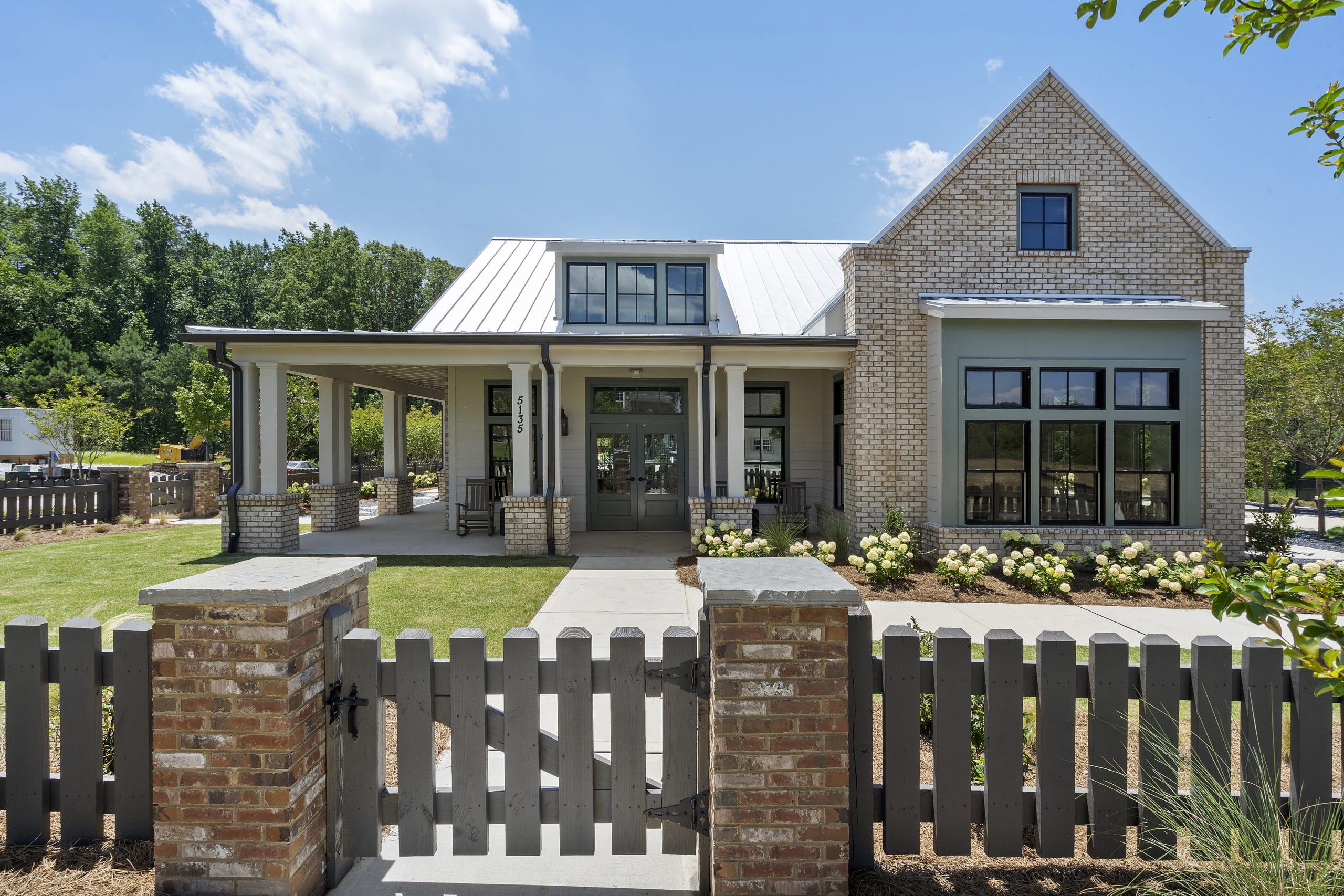 Modern house with brick and siding exterior, large windows, metal roof, covered front porch with rocking chairs, well-maintained lawn, landscaping, wooden fence with brick pillars.