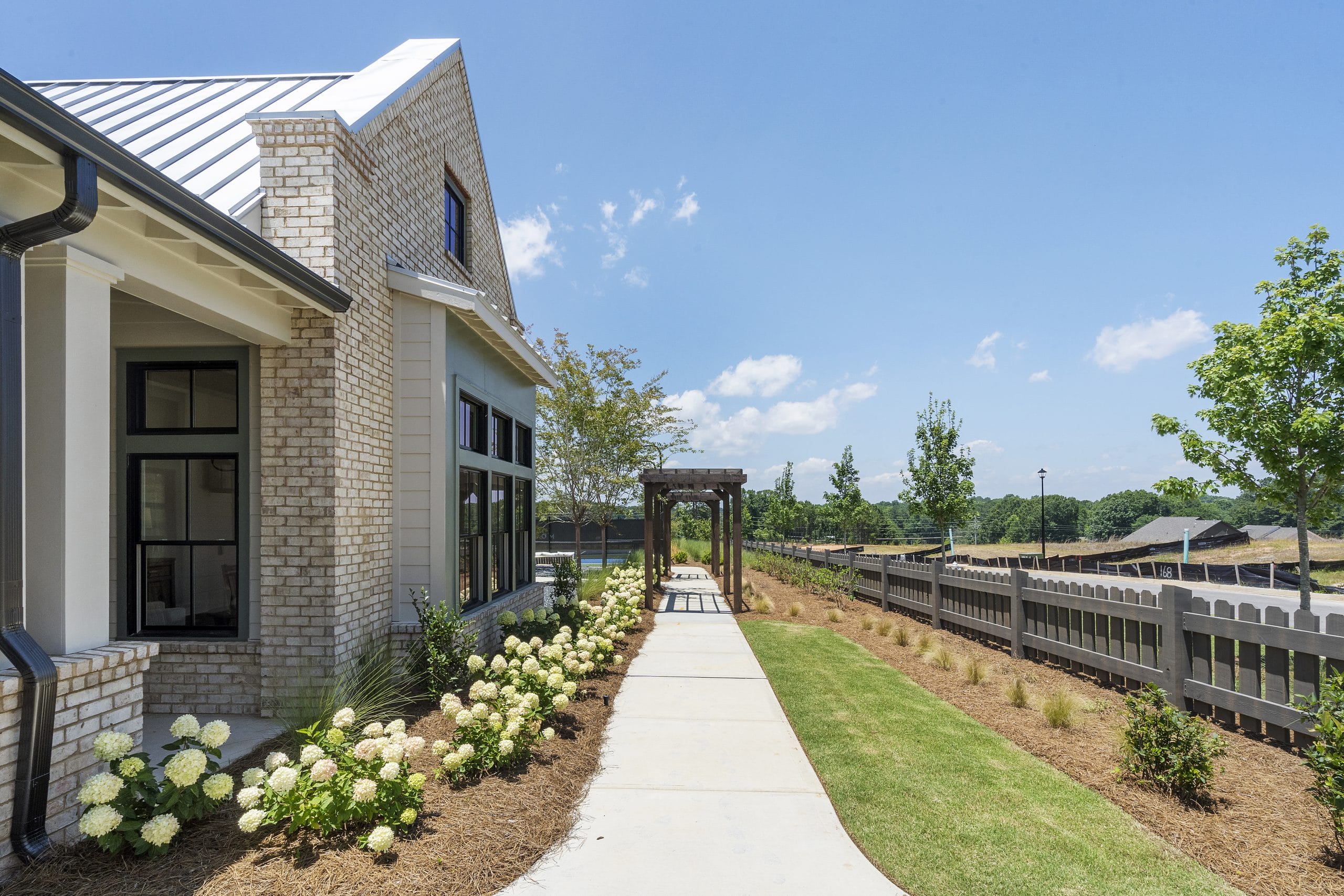 Exterior of modern house with light-colored brick walls, large windows, concrete pathway lined with white flowering plants and well-maintained lawn, wooden pergola structure, clear sky with scattered clouds, trees, and fence in the background.